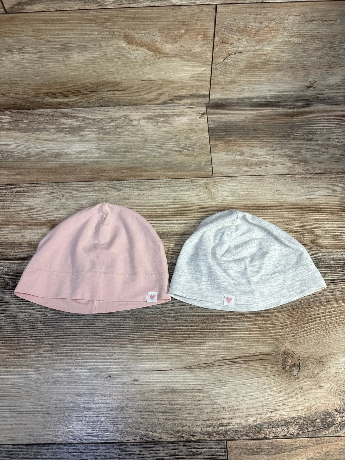 Top-down view of H&M cotton jersey beanies 2-pack grey and pink showing heart-shaped tags on a wooden floor