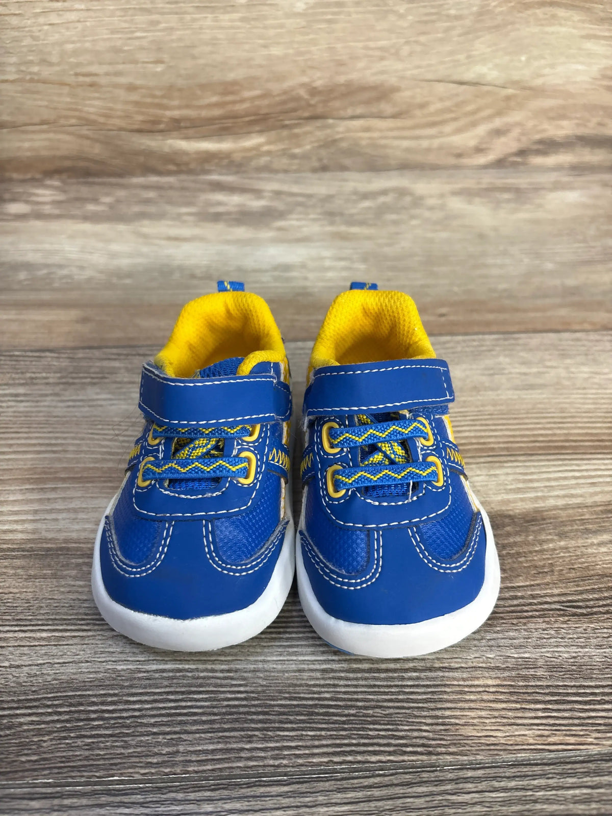 Front view of Wonder Nation blue sneakers showcasing yellow accents, hook-and-loop straps, and white cushioned soles on a wood-grain surface