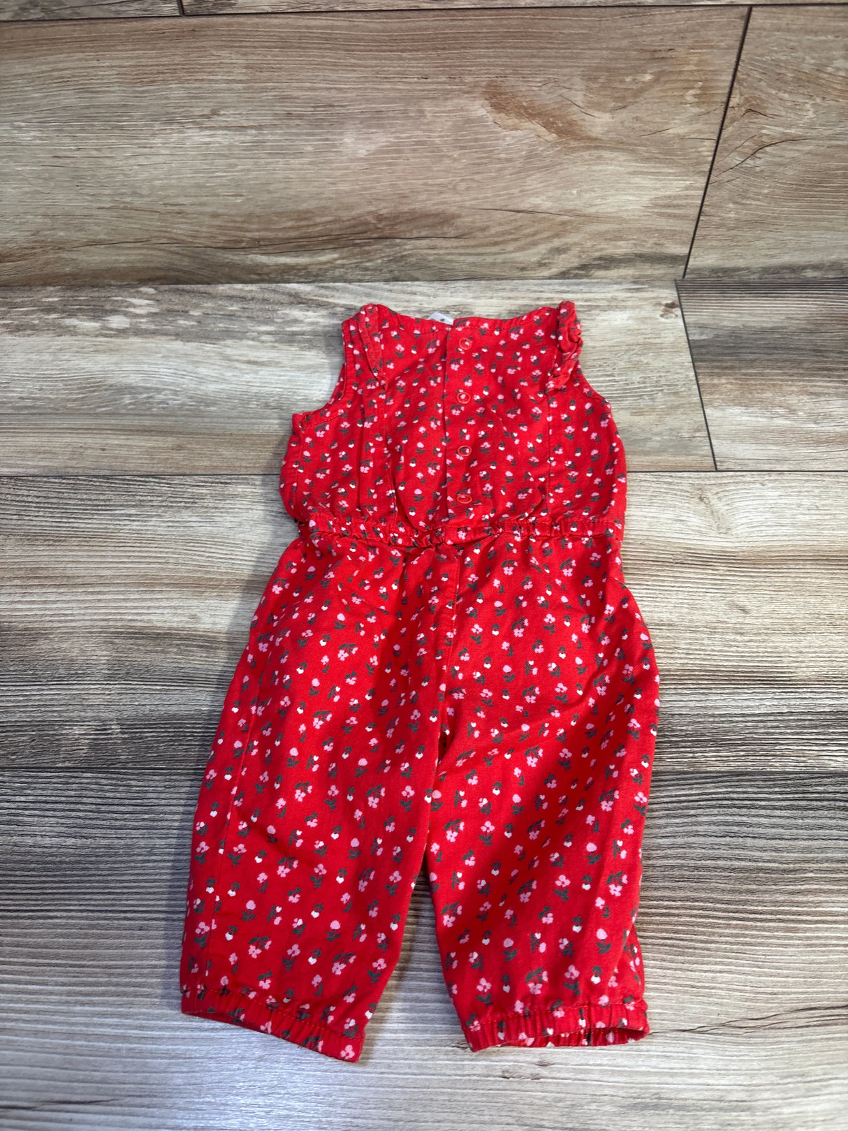 Flat lay of red floral sleeveless baby jumpsuit showing front buttons, elastic waist, and gathered leg cuffs on wood floor background