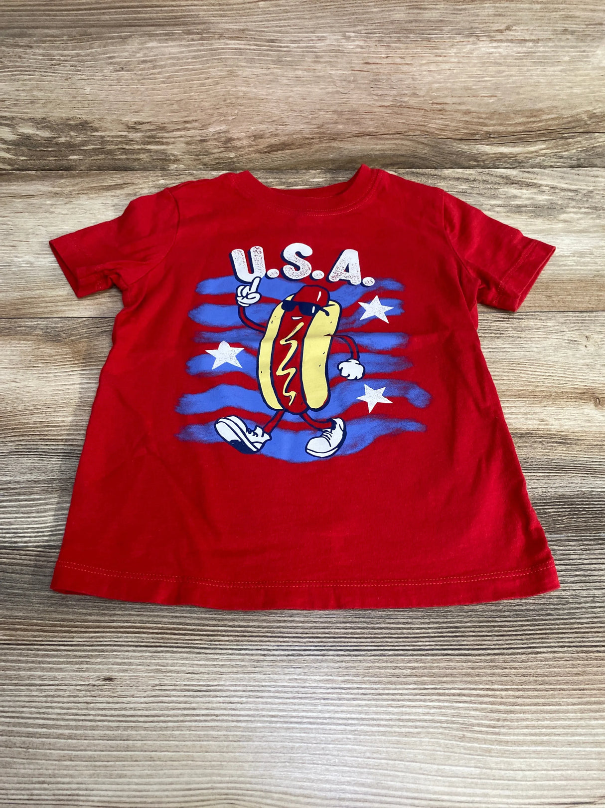 Flat lay of red OshKosh toddler t-shirt showing walking hot dog graphic with USA text and blue stars on wood background