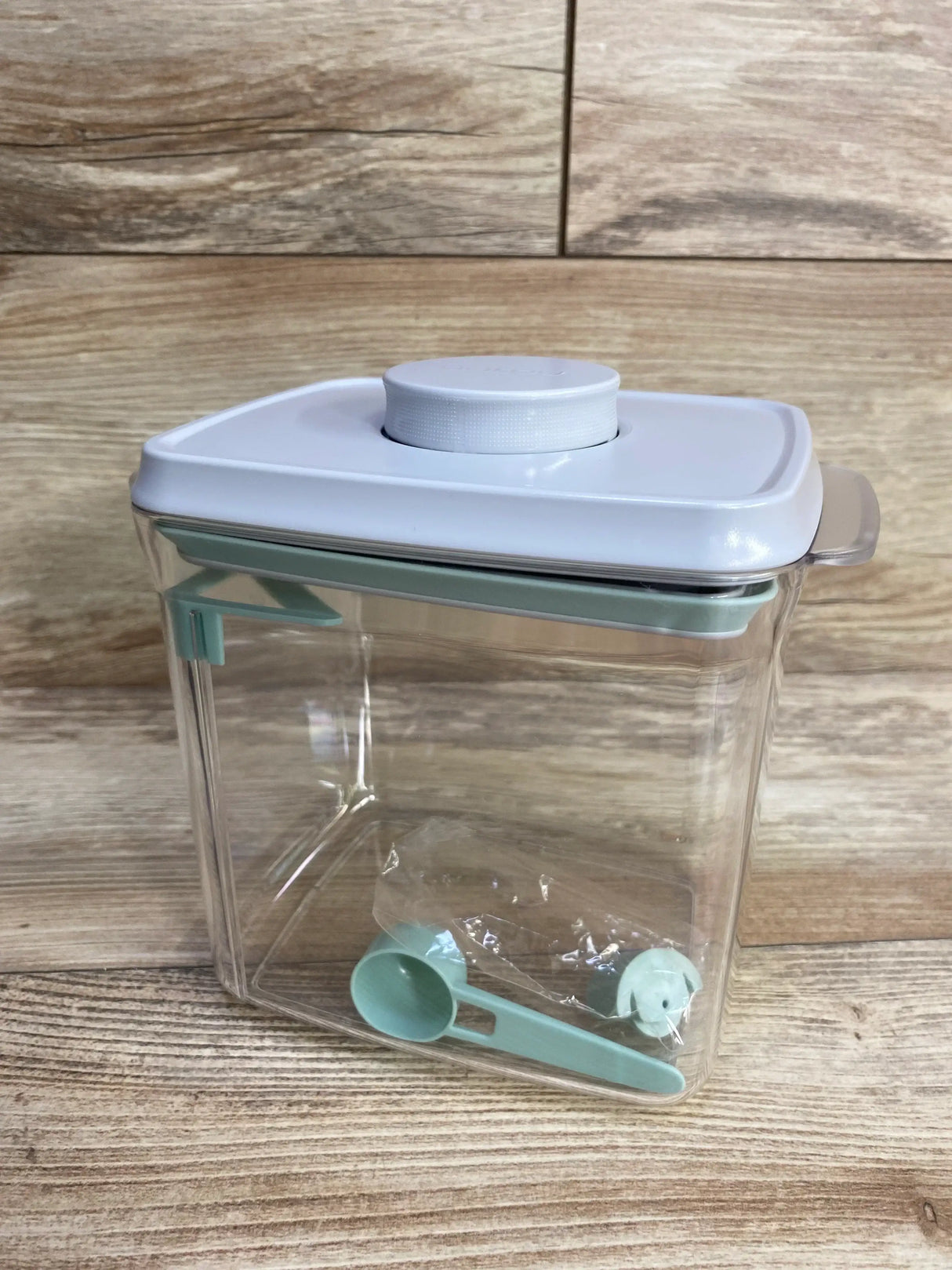 Front view of Ankou formula container showcasing clear plastic body with white airtight lid and mint scoop on wood plank floor