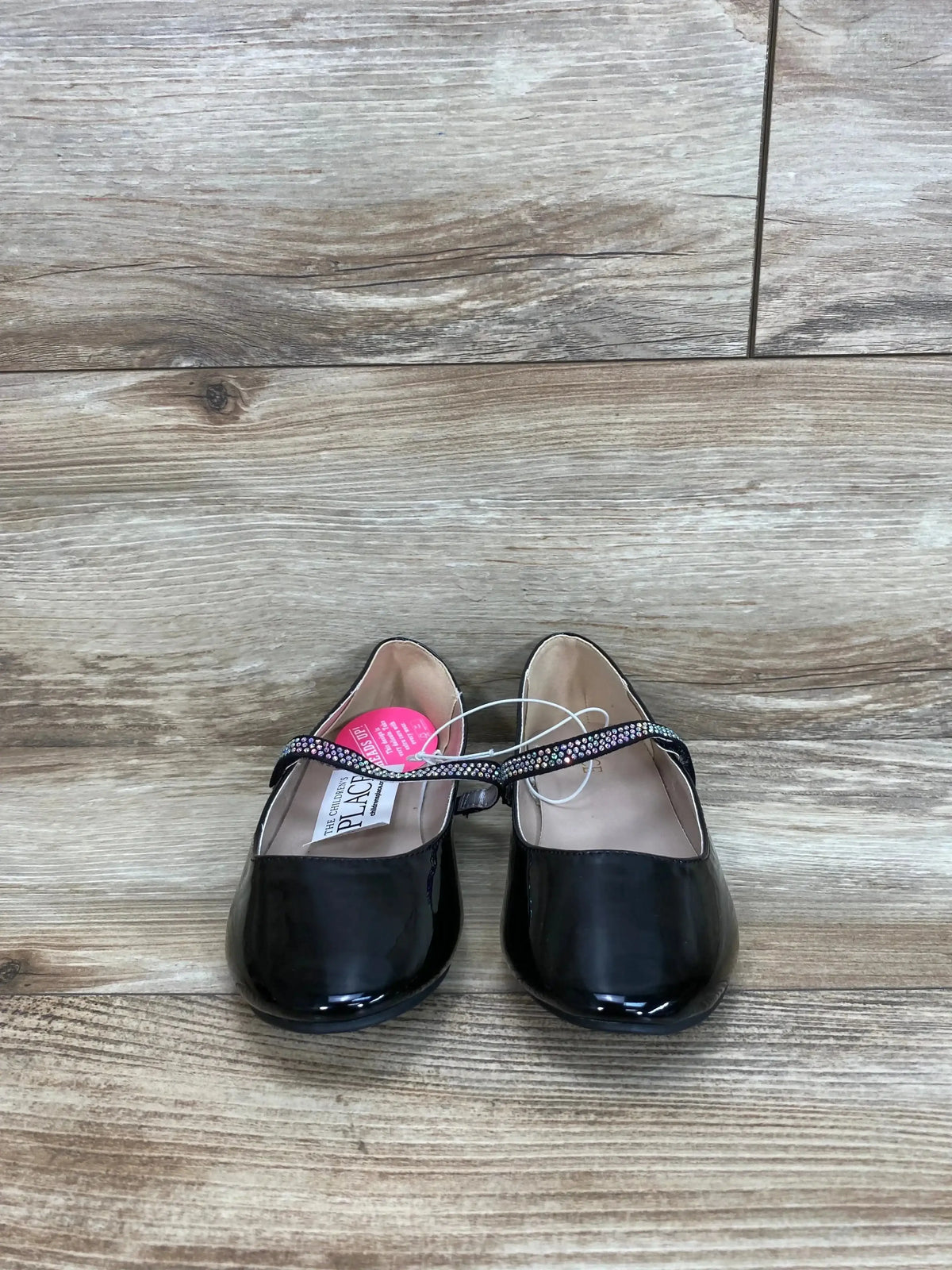 Front view of girls' black faux patent leather ballet flats showcasing jeweled Mary Jane strap on wood floor background