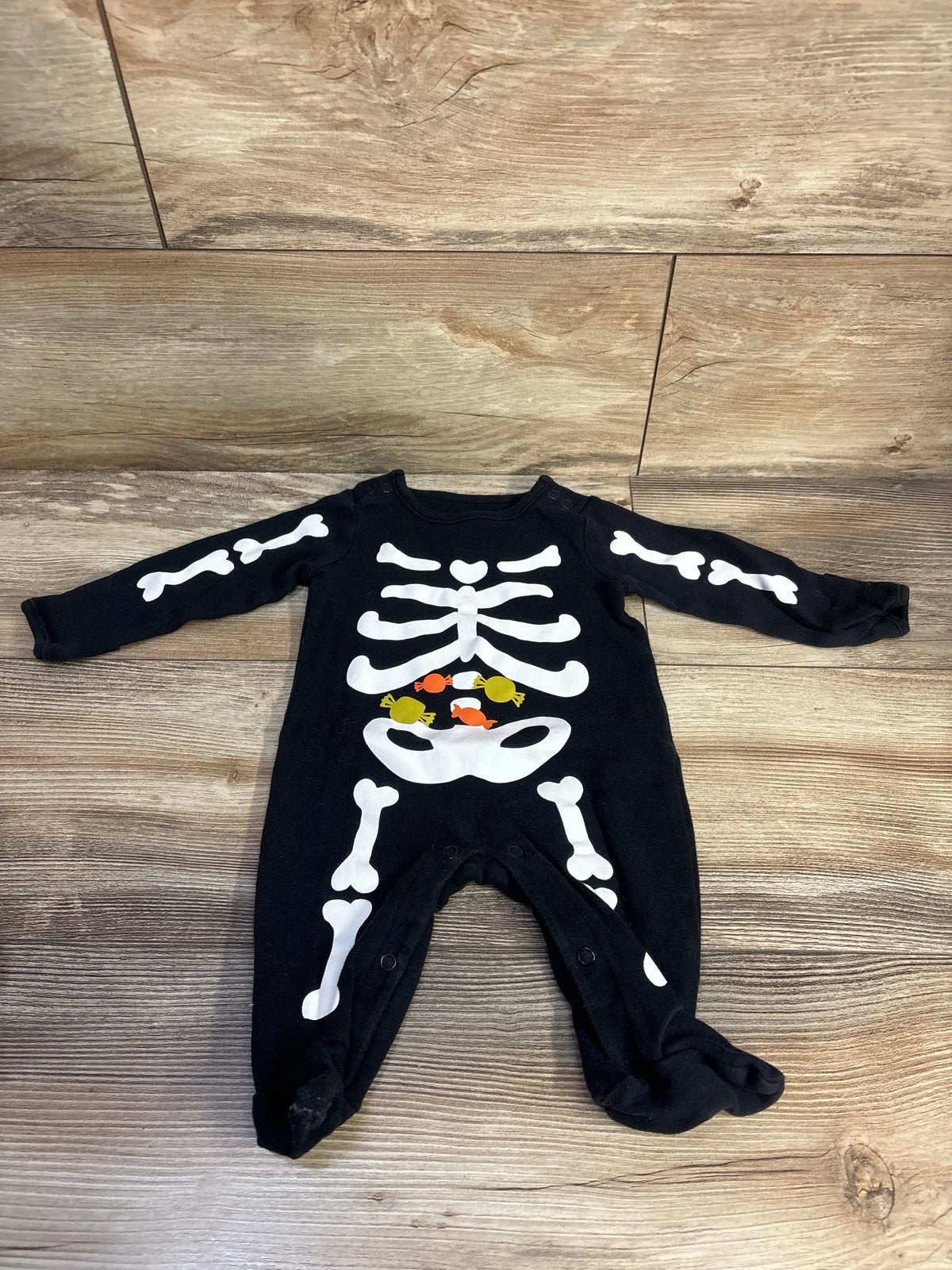 Front view of black Just One You skeleton footed coverall showcasing white skeleton print on a wood-look floor