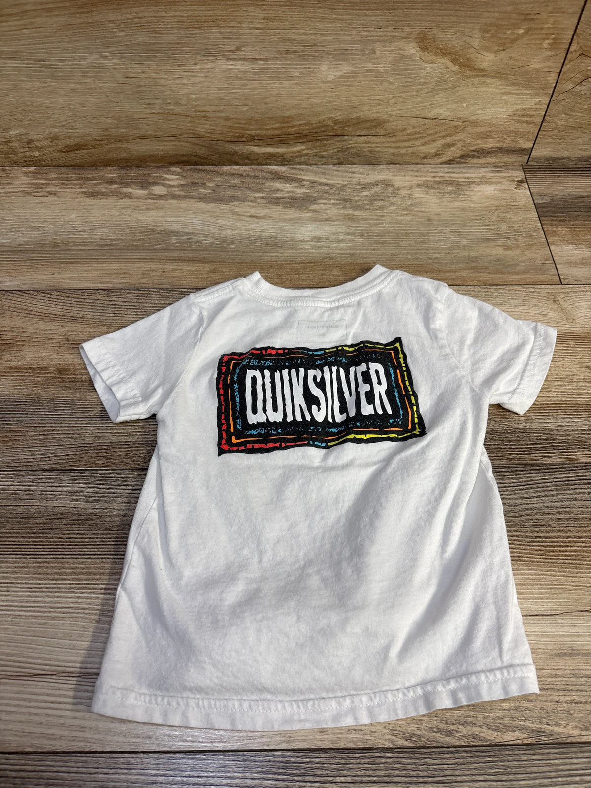 Back view of Quiksilver toddler white T-shirt showcasing multicolor framed Quiksilver logo on wood floor background