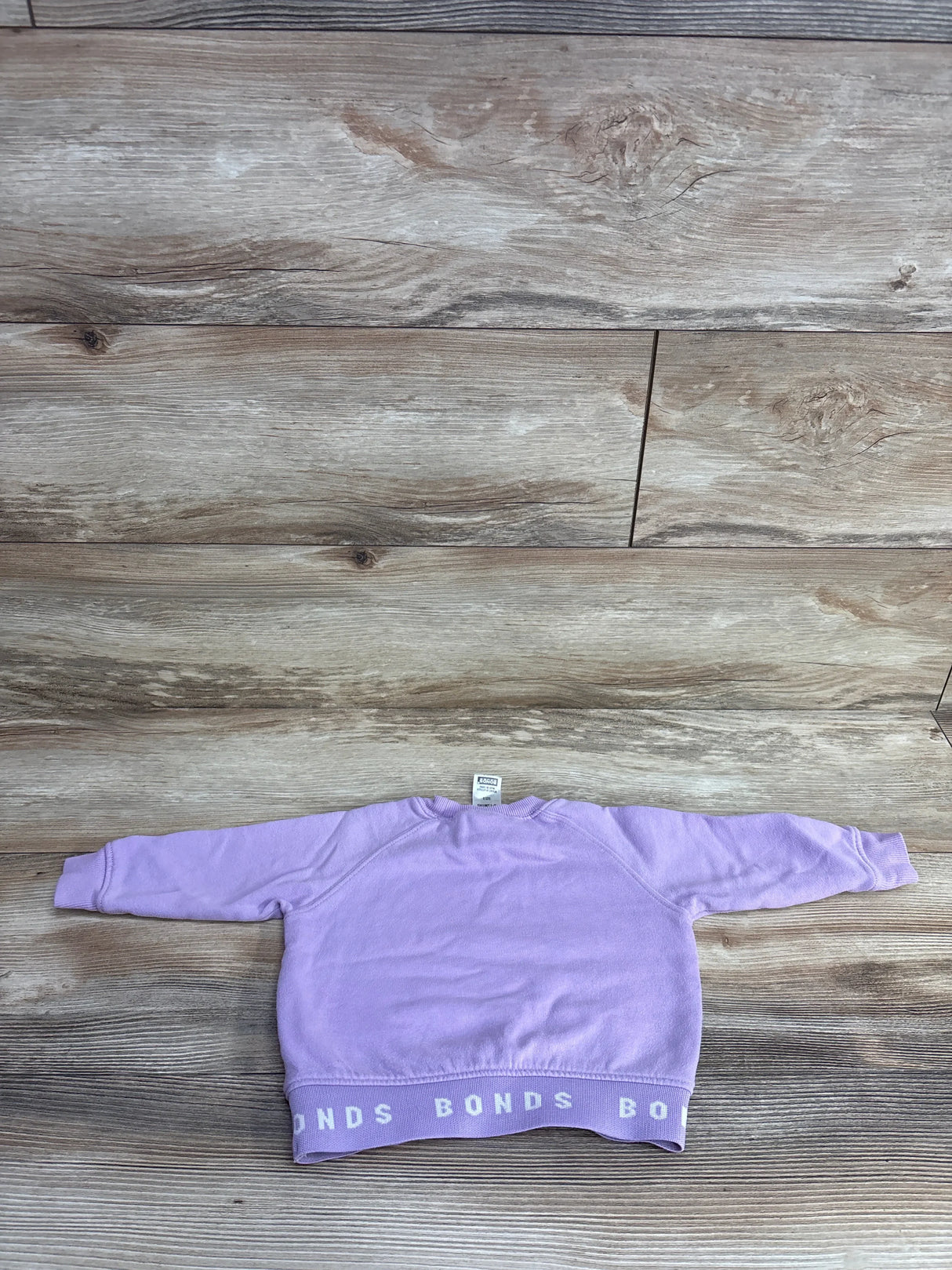 Light purple children's sweatshirt with BONDS logo band at hem laid flat on wood floor