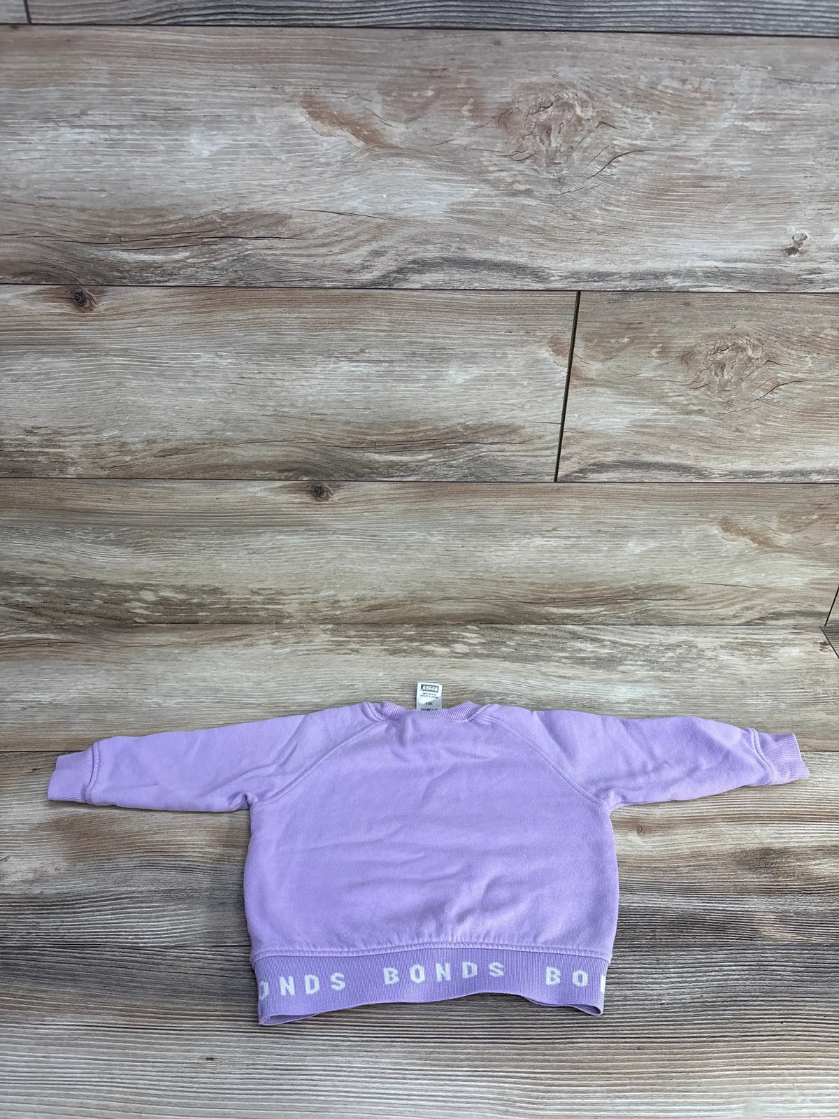 Light purple children's sweatshirt with BONDS logo band at hem laid flat on wood floor