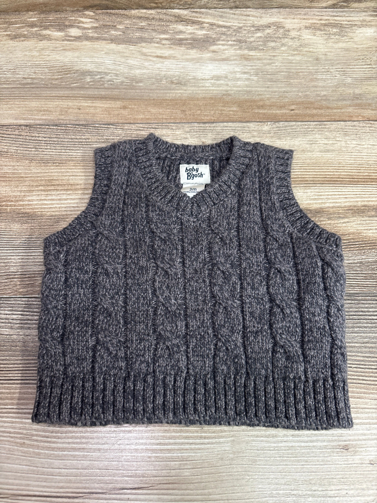 Front view of a gray Oshkosh sweater knit vest showcasing cable-knit detailing and V-neck with wood-grain background