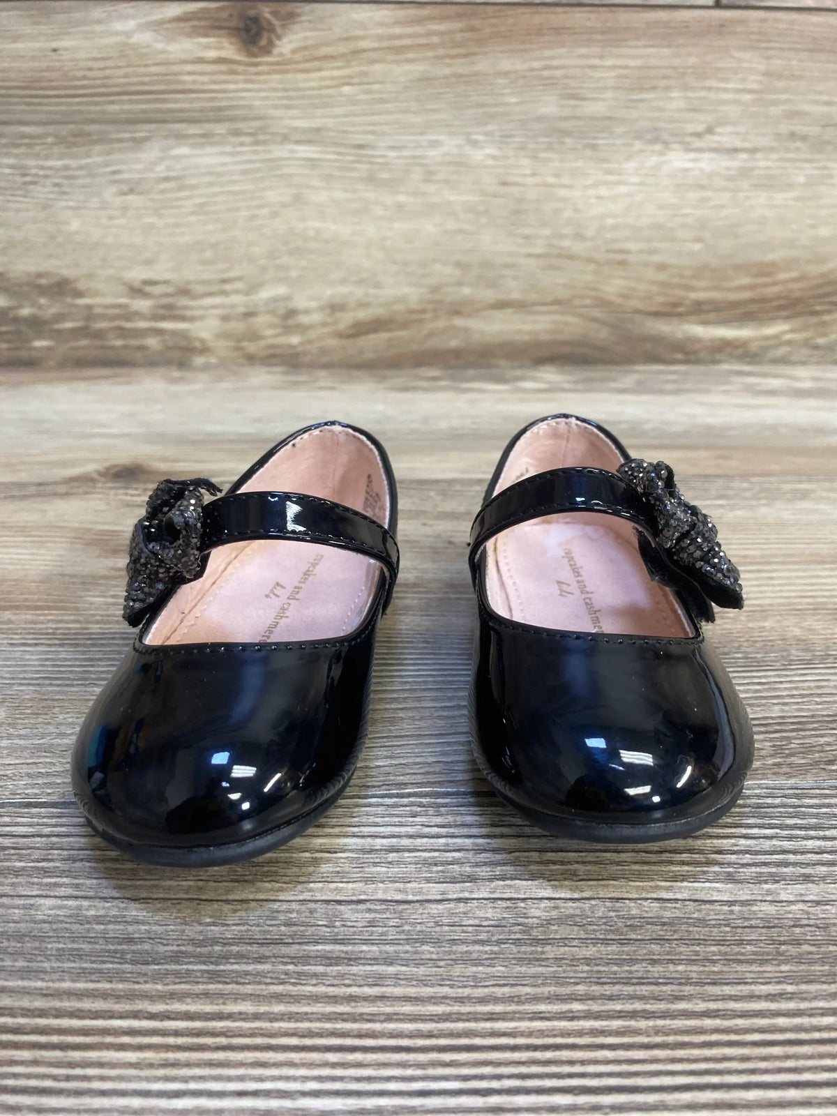 Black patent leather girls' dress shoes with bow detail on wood floor