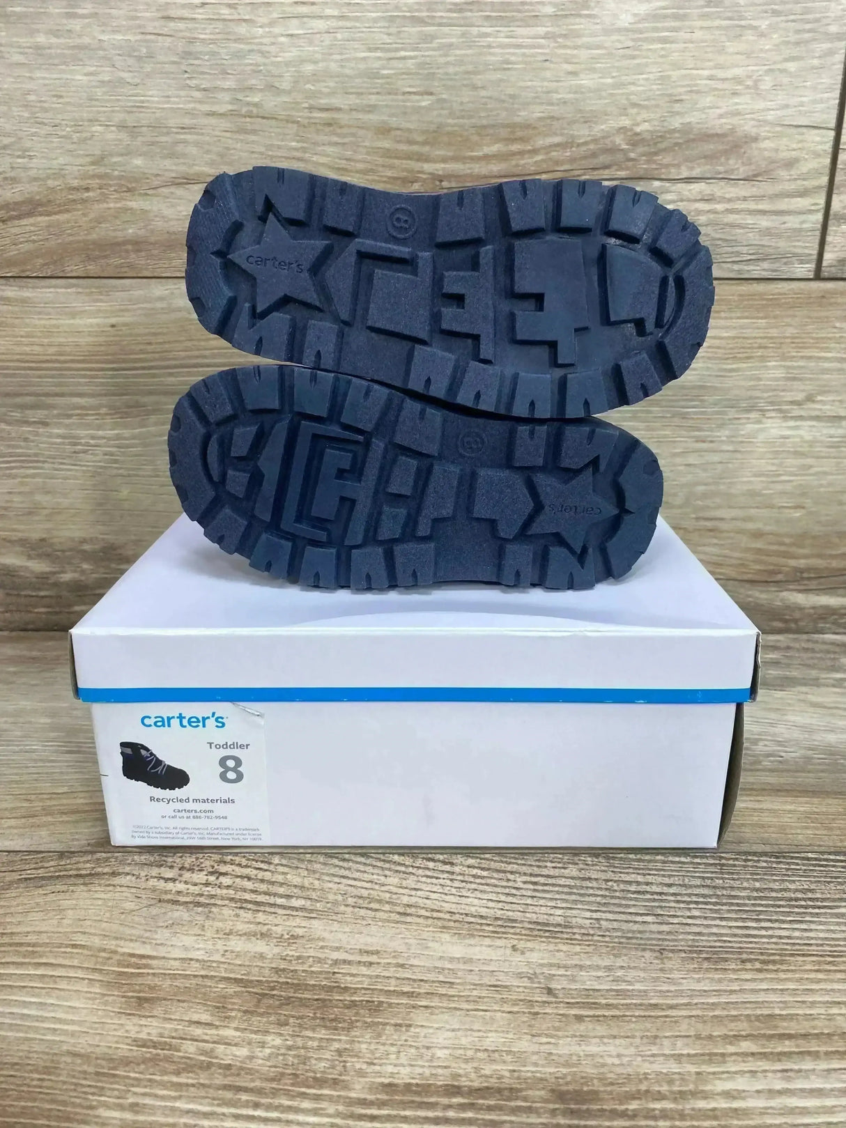 Bottom view of Carter’s toddler Fallon boots showing navy soles with star logo and deep treads on white box over wood floor