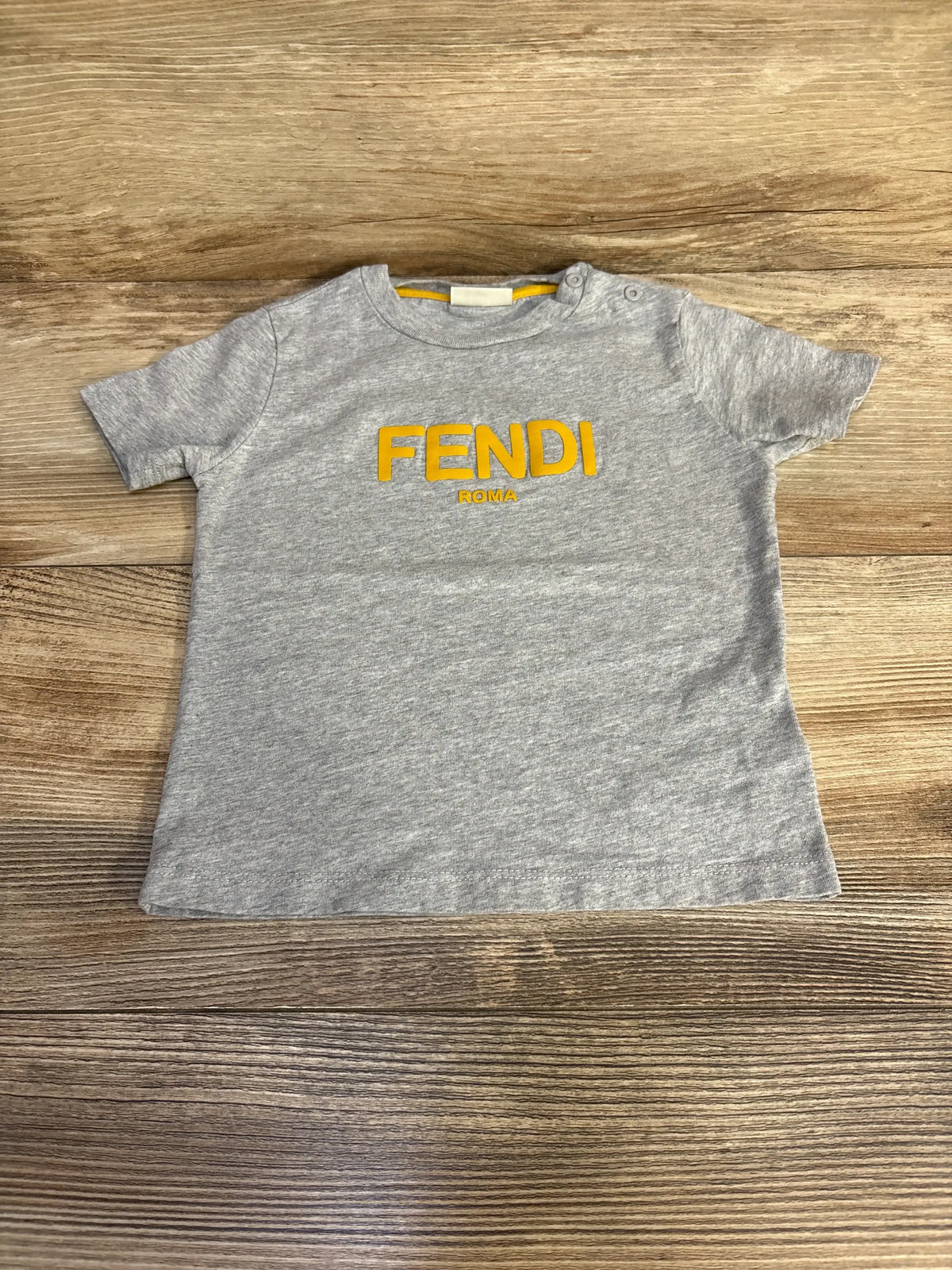 Flat lay of grey Fendi toddler T-shirt (18 months) showcasing yellow FENDI ROMA logo on front with wood plank background