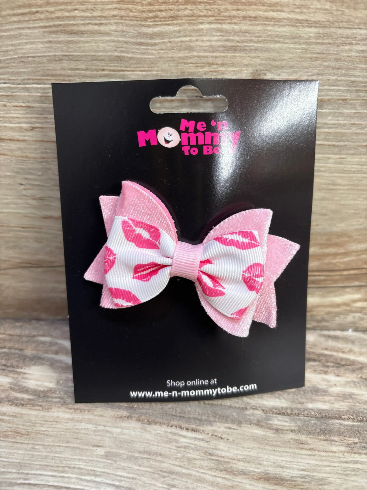 Front view of kiss-print velvet hair bow with pink lip-print grosgrain ribbon layered on pink velvet, on black display card branded Me n Mommy To Be