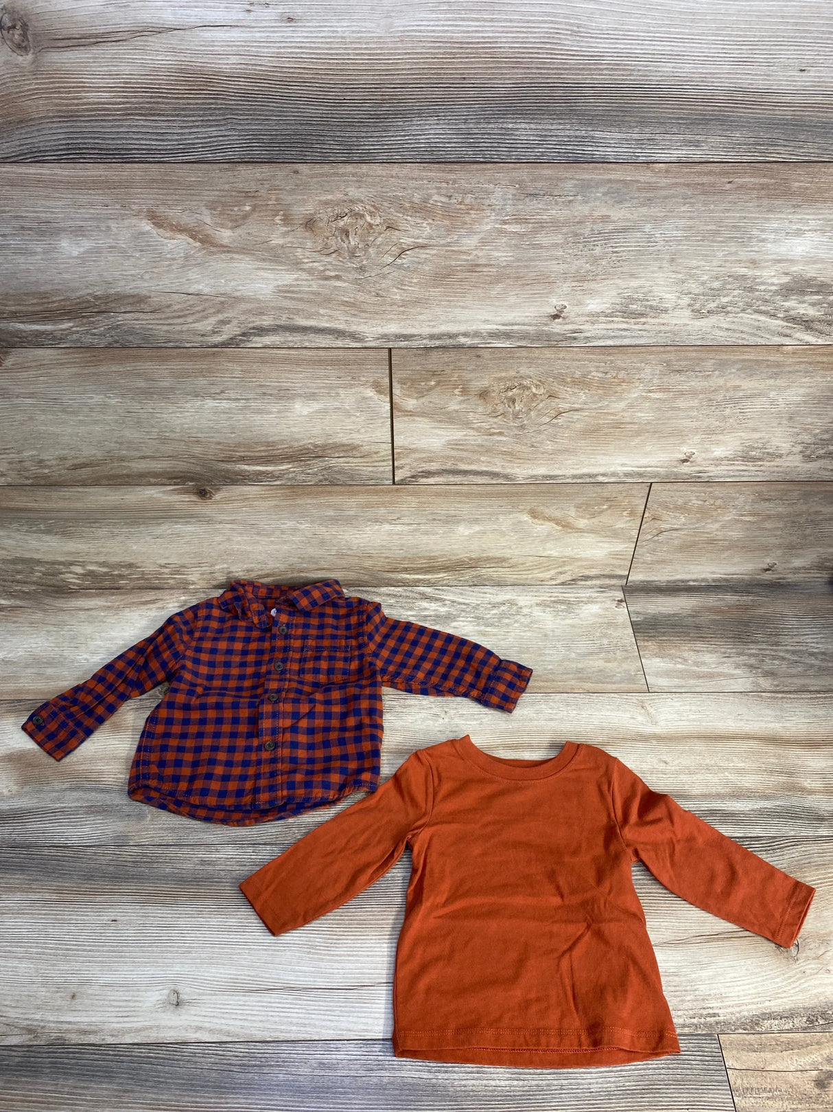 Orange and blue checkered baby shirt with buttons and solid orange long-sleeve baby tee on wood floor