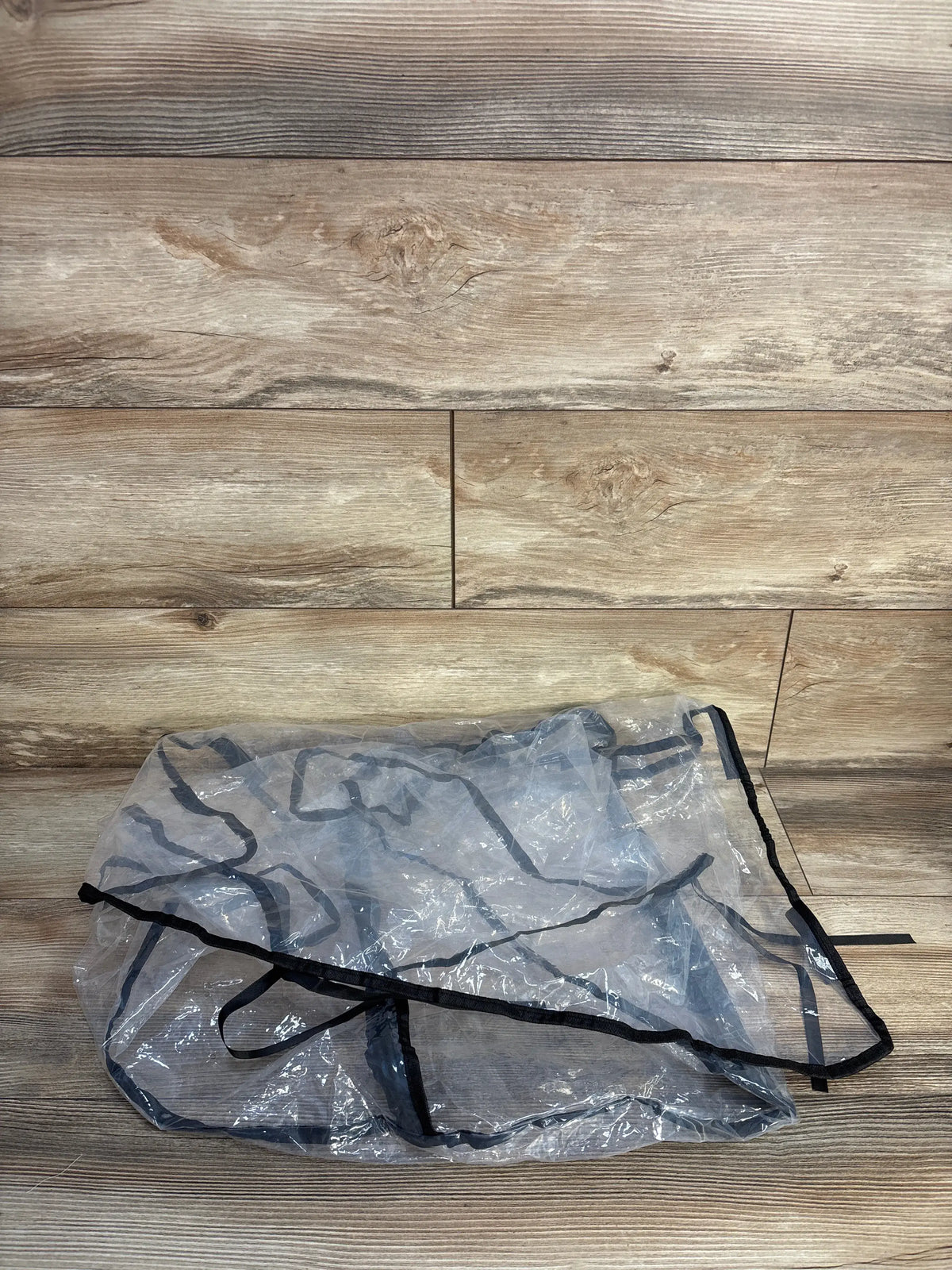 Flat lay of transparent stroller and car seat rain cover showing black trim and tie straps on wood background