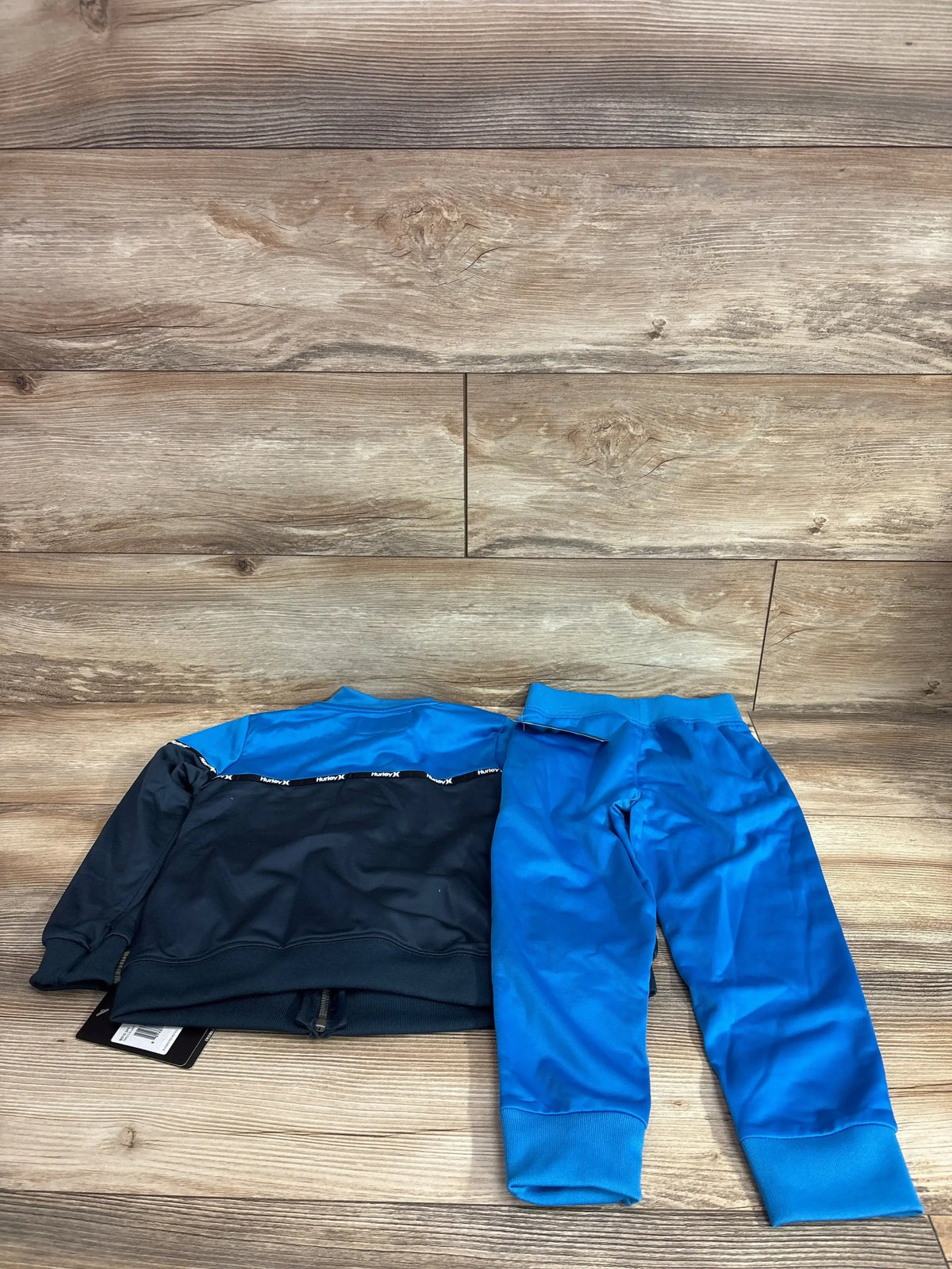 Flat lay of blue Hurley toddler 2-piece tricot tracksuit showing zip jacket with contrast yoke and elastic-waist pants on wood background