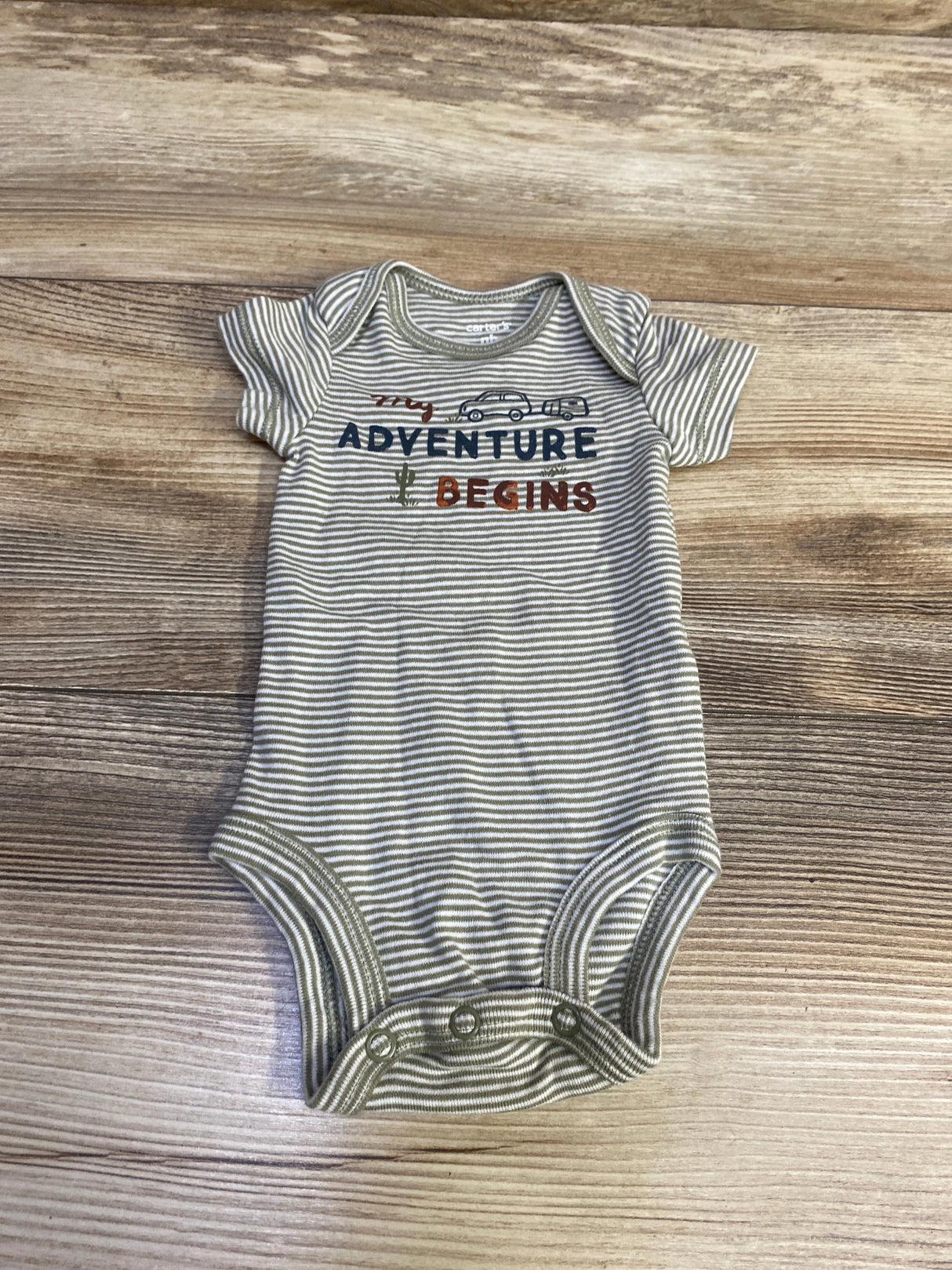 Flat lay of newborn green striped bodysuit with My Adventure Begins text, car and cactus graphics on wood