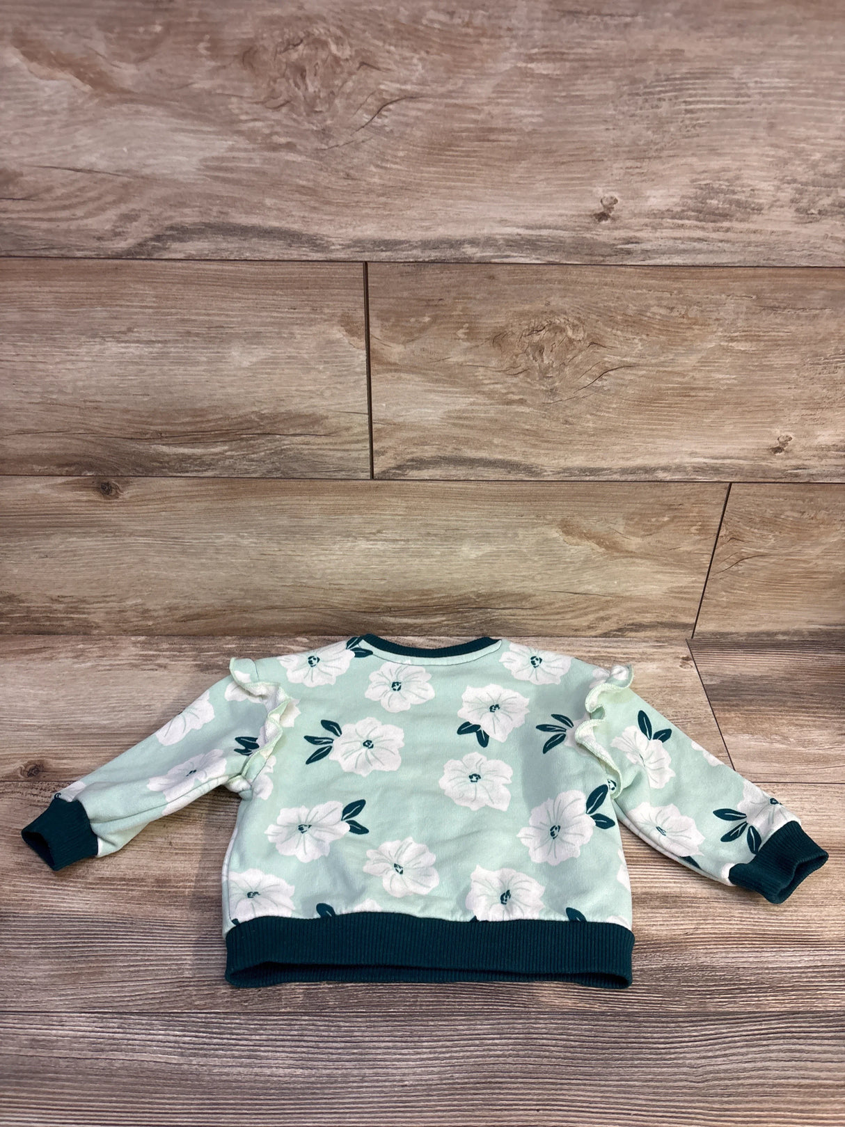 Flat-lay of mint green toddler pullover showcasing white floral print, ruffle shoulders, and dark-teal ribbed cuffs on wood