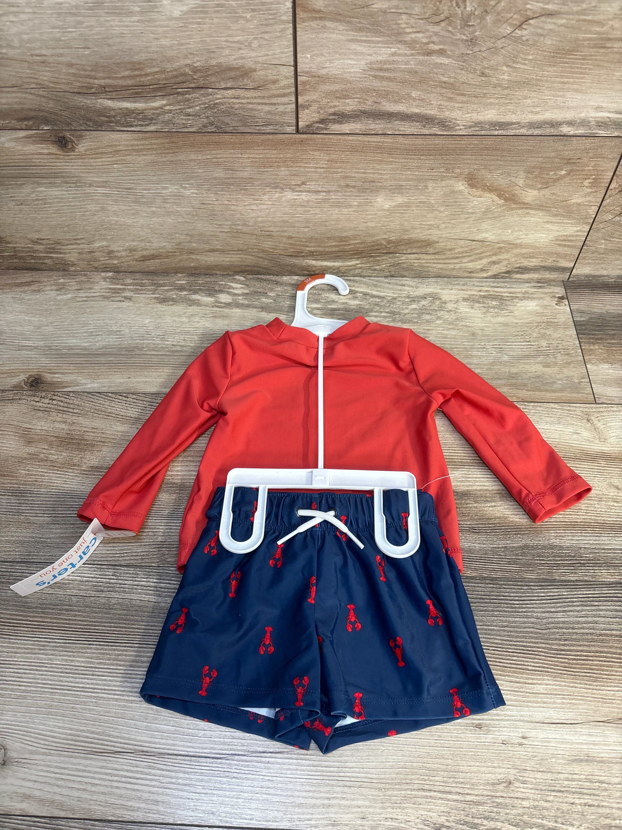 Toddler boy's red rash guard and navy swim shorts with red lobster print on wood background