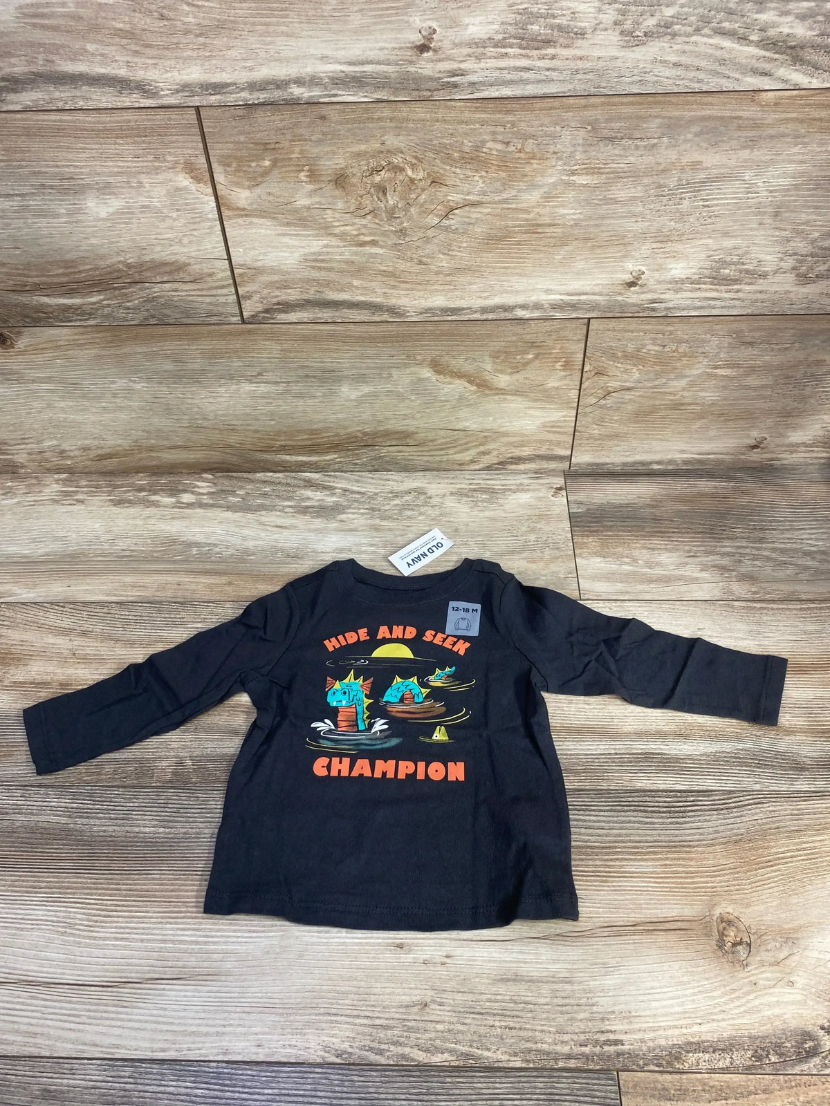 Front view of Old Navy toddler long-sleeve grey shirt showing Hide and Seek Champion text and dinosaur print on wooden floor