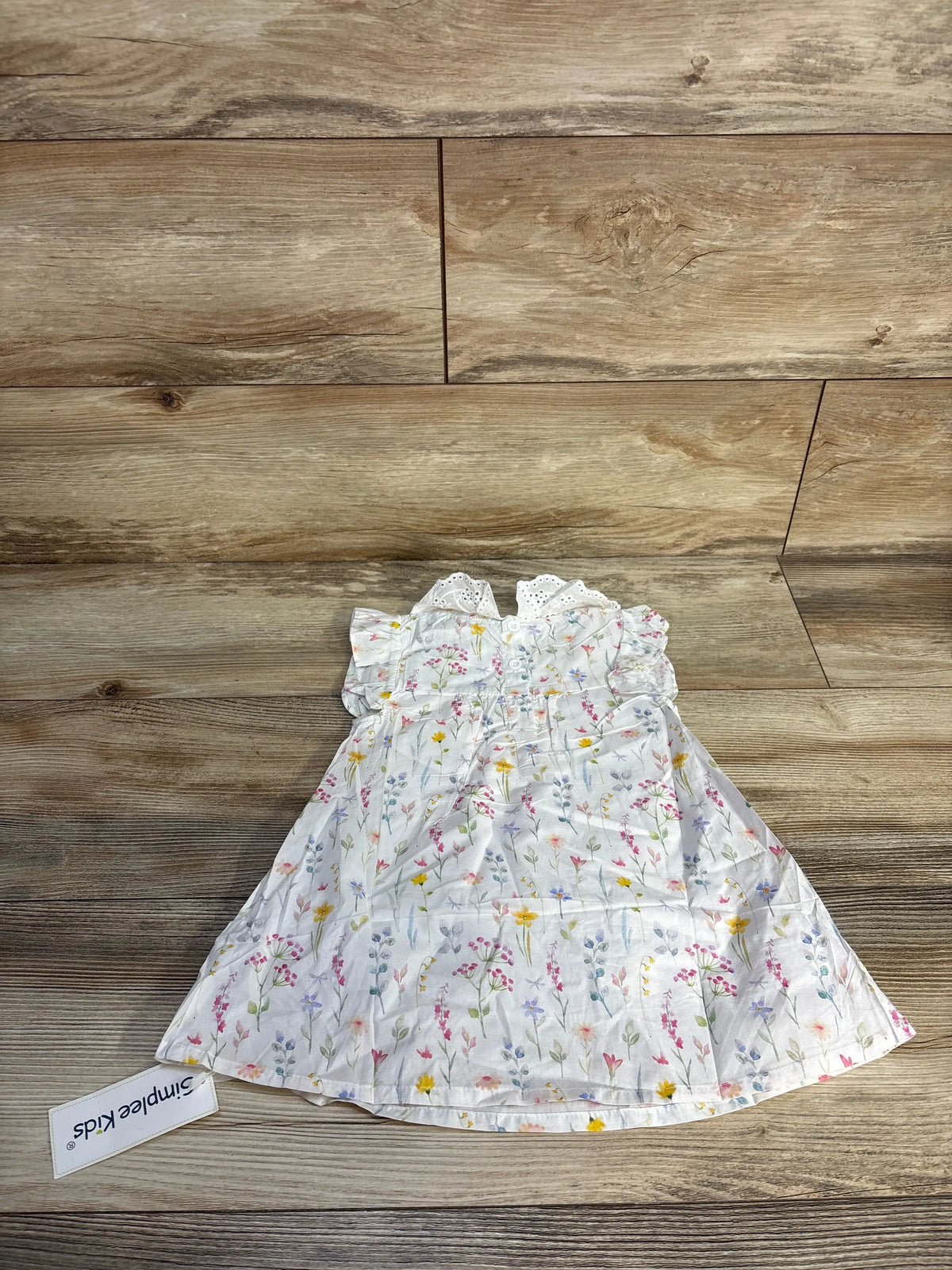 Front view of white SimpleKids floral eyelet collar dress for ages 6 to 9 months, showcasing eyelet collar on wood background
