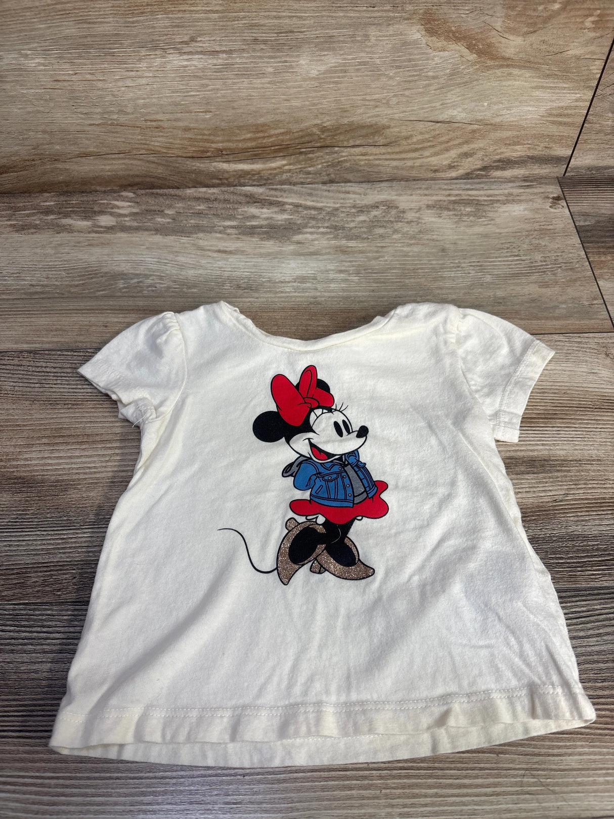 Front view of ivory Baby Gap Minnie Mouse toddler shirt with centered Minnie graphic, red bow, glitter shoes on wood