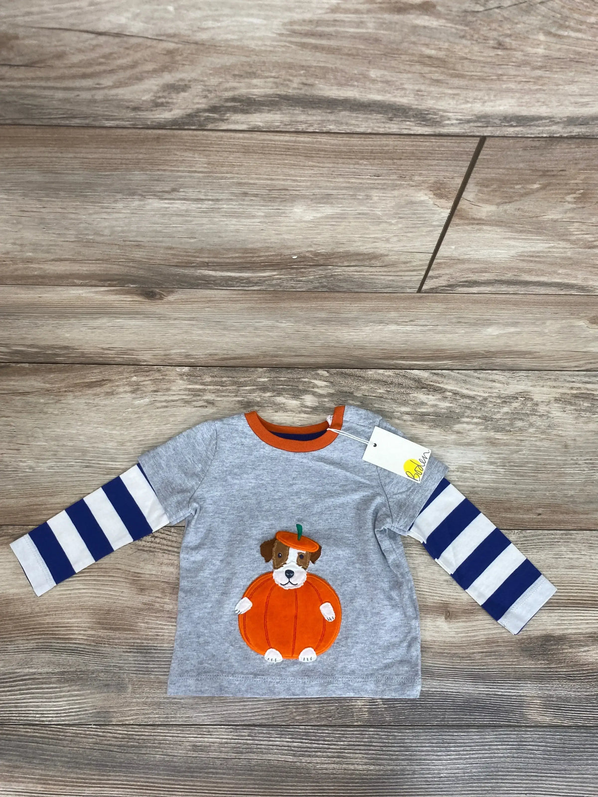 Front view of a Baby Boden 2fer puppy pumpkin shirt with striped sleeves on a wood grain background