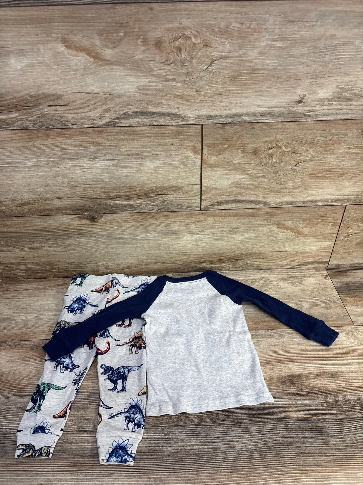 Top view of toddler dinosaur 2-piece pajamas: grey raglan top with navy sleeves and grey dinosaur-print pants on wood floor