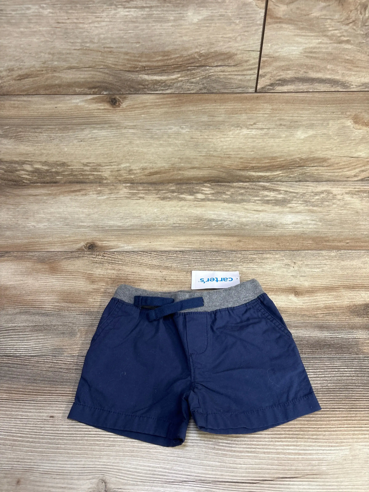 Top-down view of navy Carter’s baby shorts size 6M showing gray elastic drawstring waistband on wood background