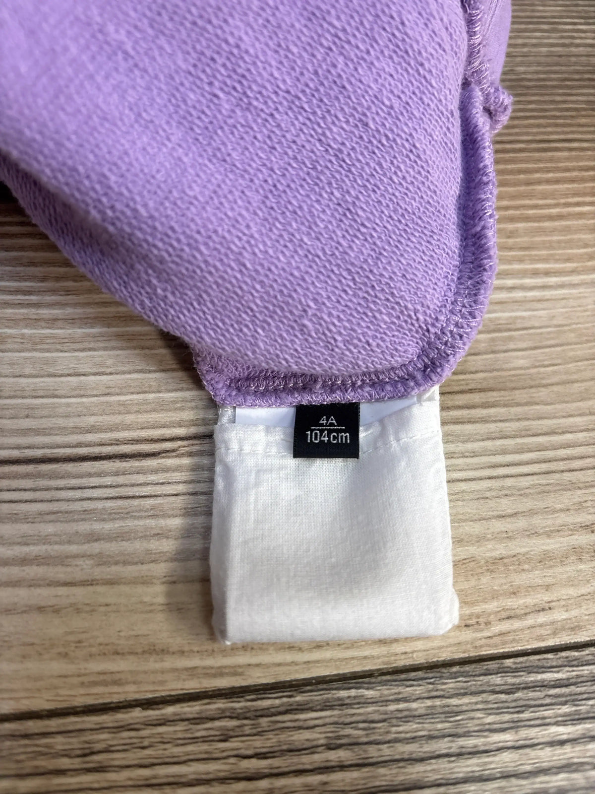 Close-up of Lilac Girls Versace Medusa hoodie showing size tag 4A 104cm on white care label with wooden background