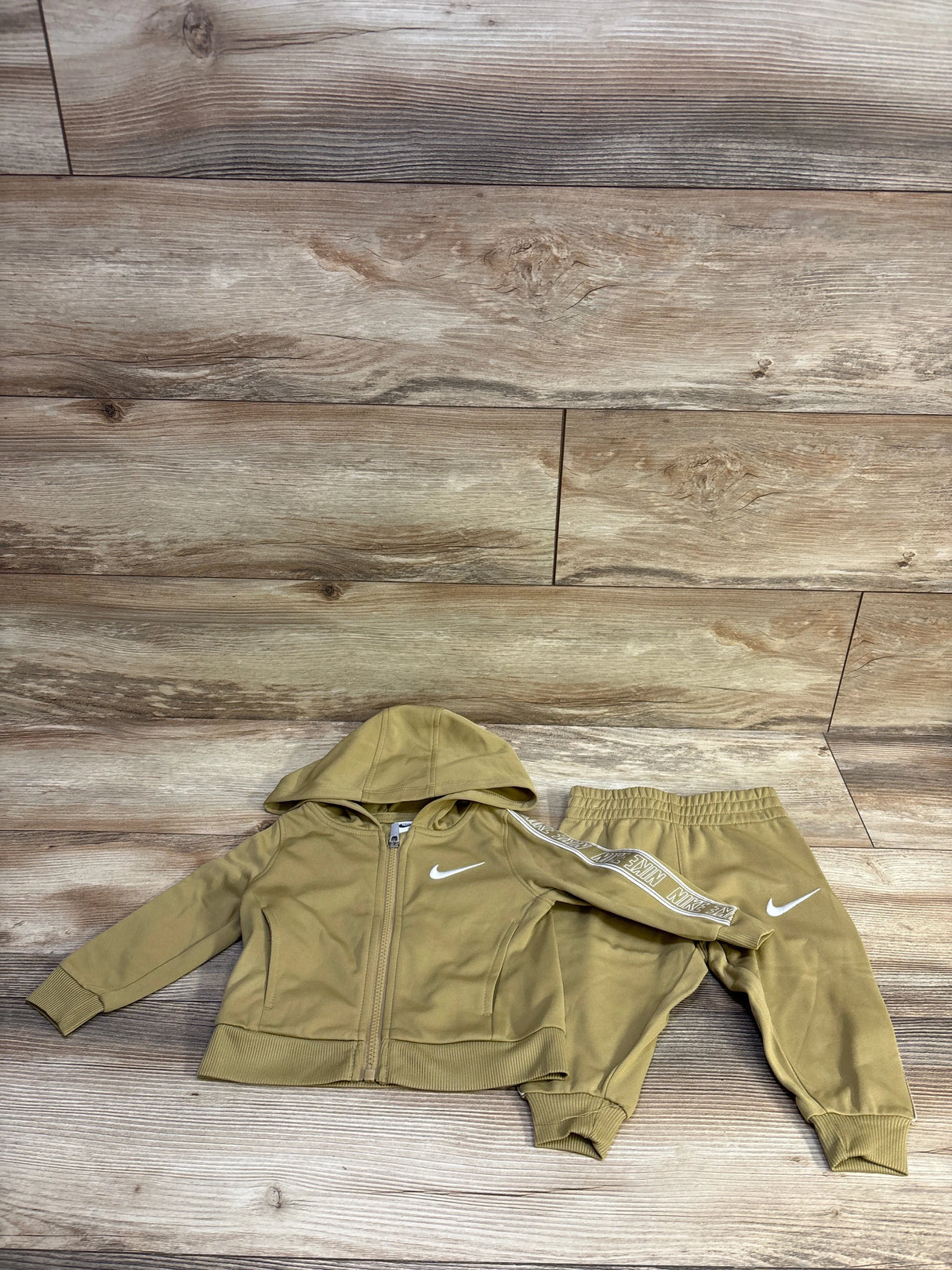 Flat lay of tan Nike Dri-Fit 2-piece tracksuit (12M) showing zip hoodie with white swoosh and sleeve logo tape, elastic pants on wood background