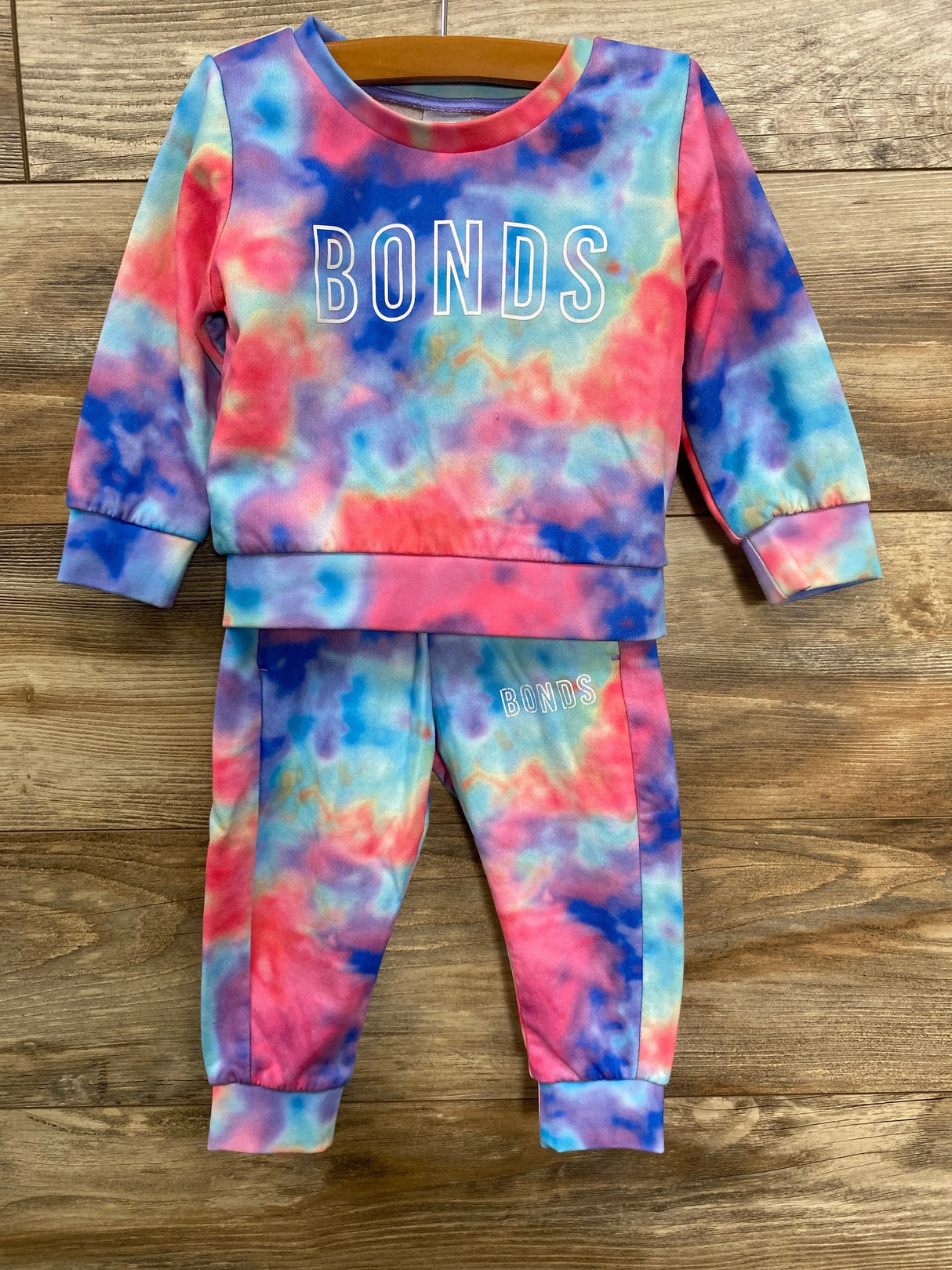 Colorful tie-dye kids' sweatshirt and pants set with BONDS logo, displayed on wooden background