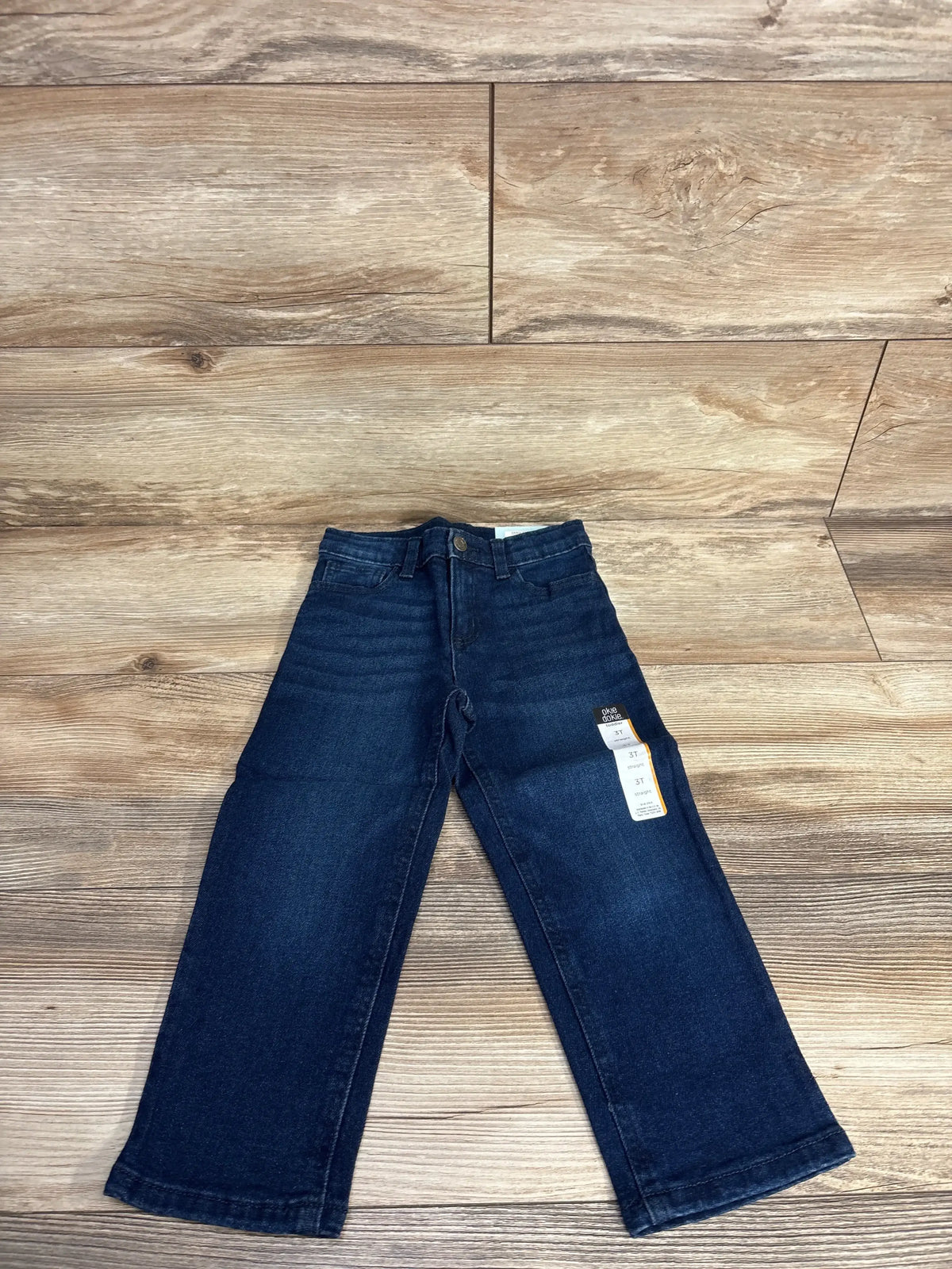 Flat lay of Okie Dokie navy toddler jeans (3T) showing original tags and slight bootcut legs on wood floor background