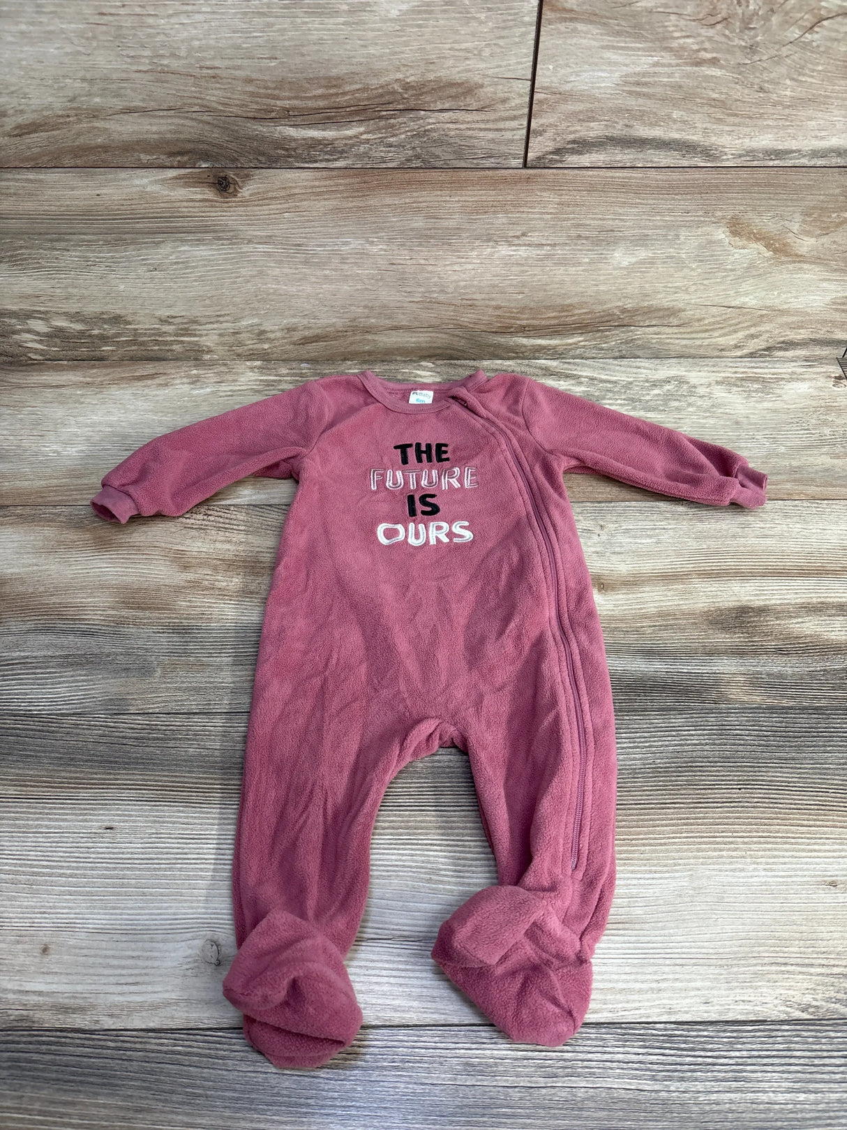 Pink fleece baby onesie with footies and zipper, embroidered with 'The Future Is Ours' on front, laid on wooden floor