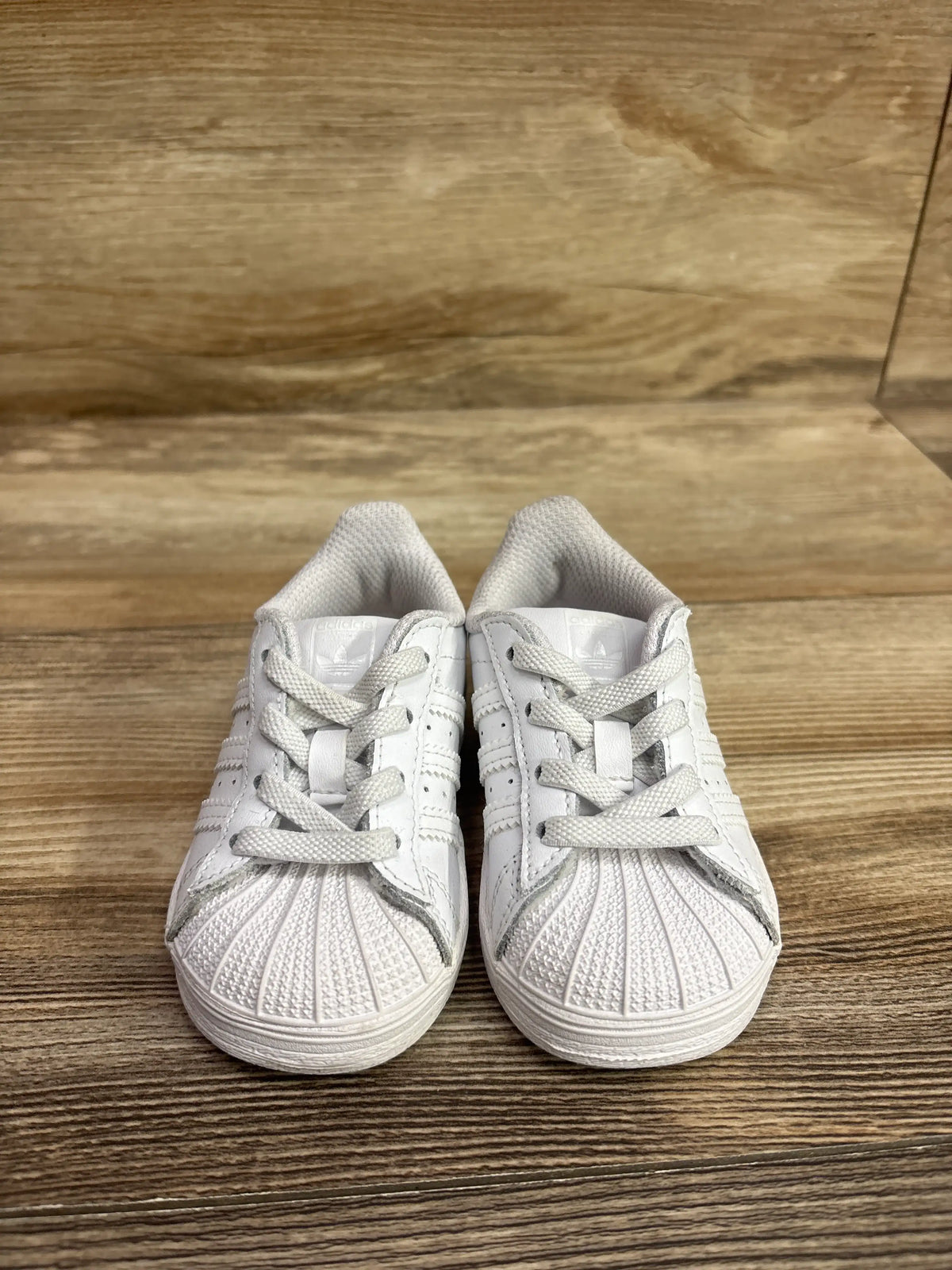 Front view of Adidas Superstar El I sneakers in white, size 5C, showcasing shell toes and laces on a wood surface