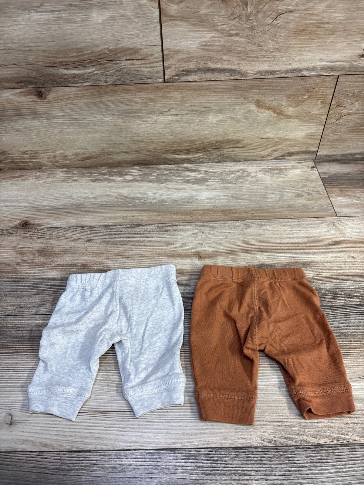 Flat lay of two newborn Carter’s pants, light gray and rust brown, with elastic waists and cuffed legs on wood background