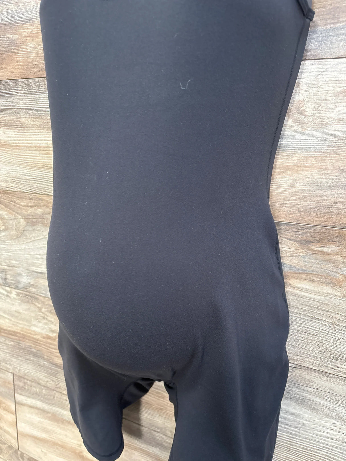 Close-up front view of Isabel Maternity Active Romper in black showing stretchy high rise maternity belly panel and mid thigh shorts on wood floor background