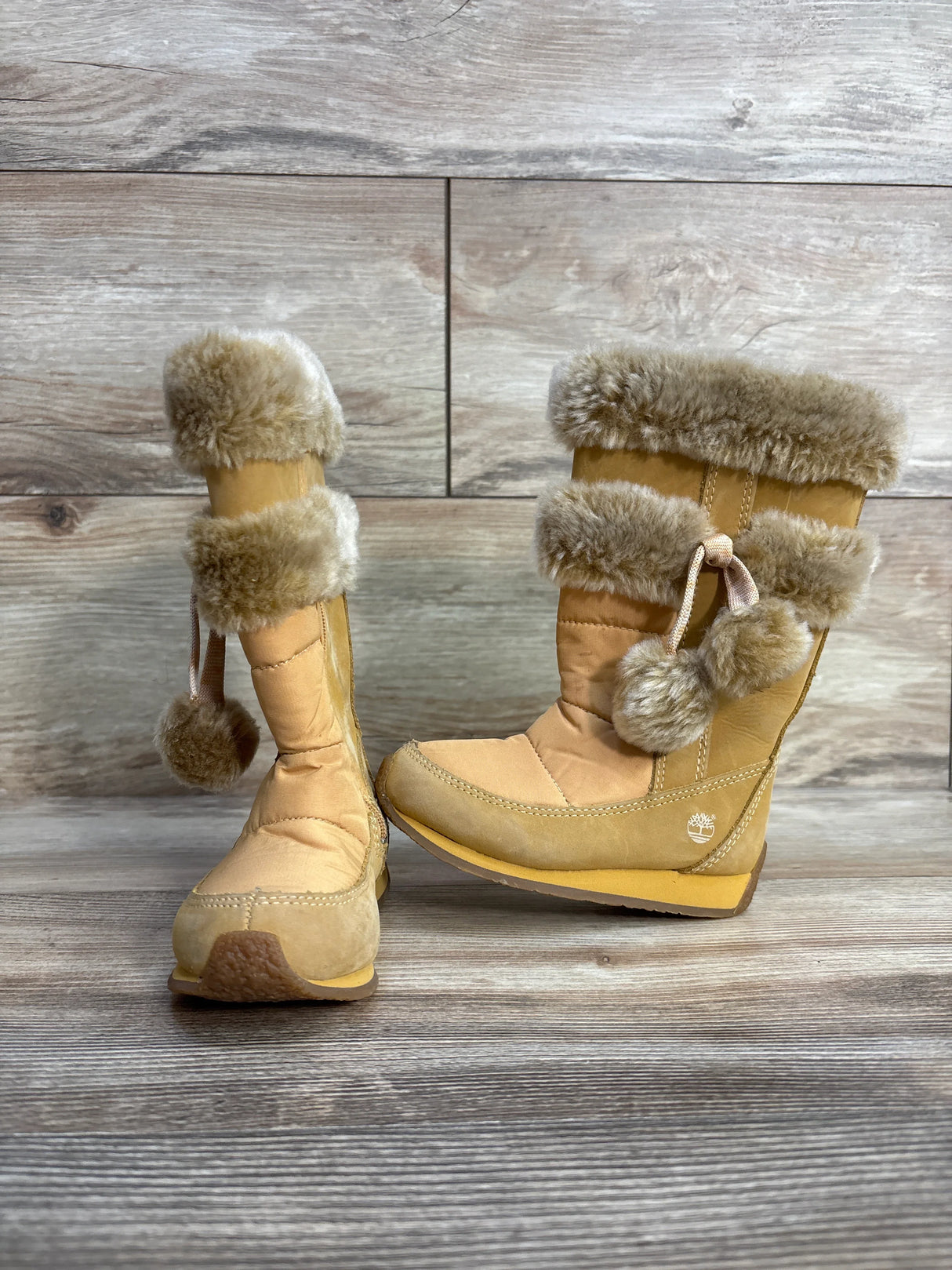 Tan Timberland winter boots with faux fur trim and pom-poms on wood floor background
