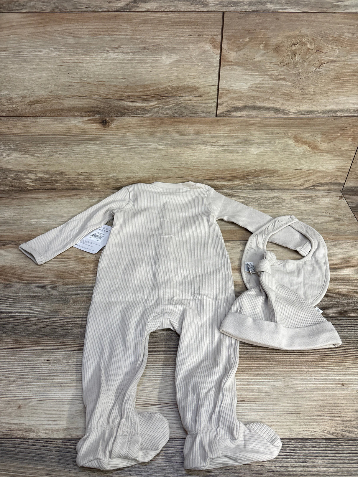 Neutral ivory ribbed baby footed onesie, matching hat, and bib set on wooden floor backdrop