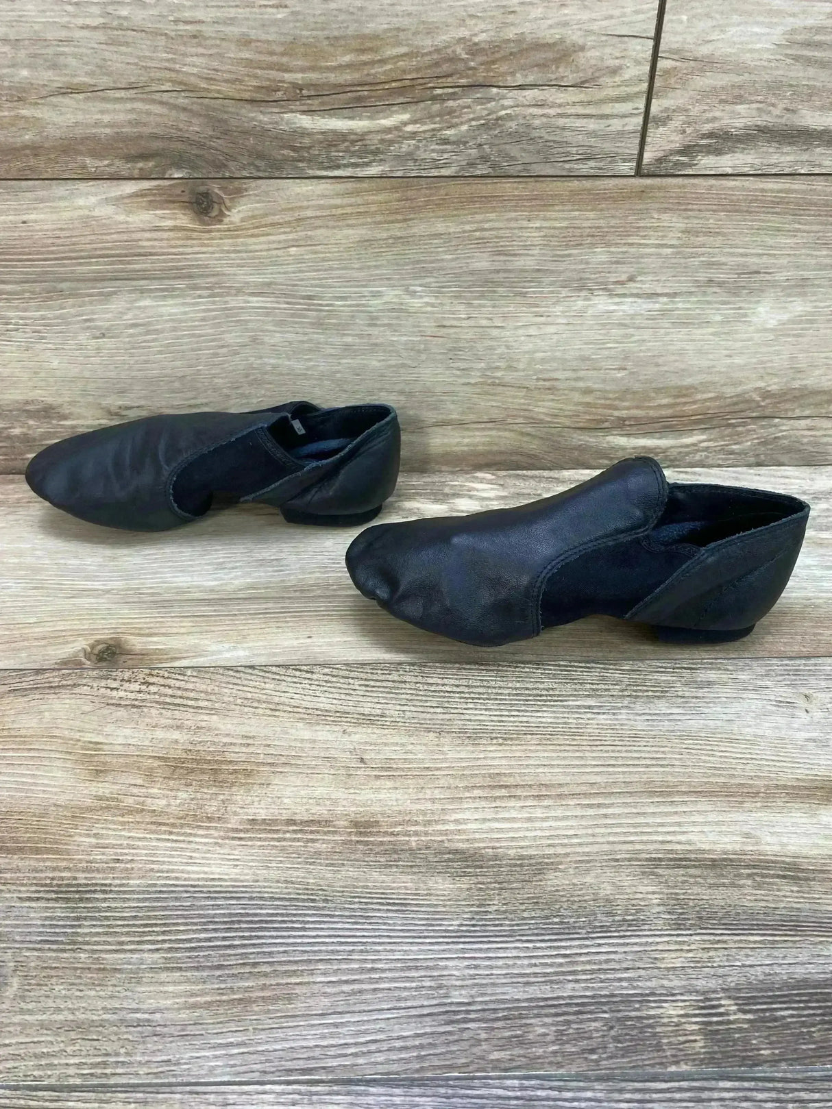 Front-side view of Capezio E-Series black jazz slip-on shoes, child size 8C, showing low heel and elastic panels on wood floor