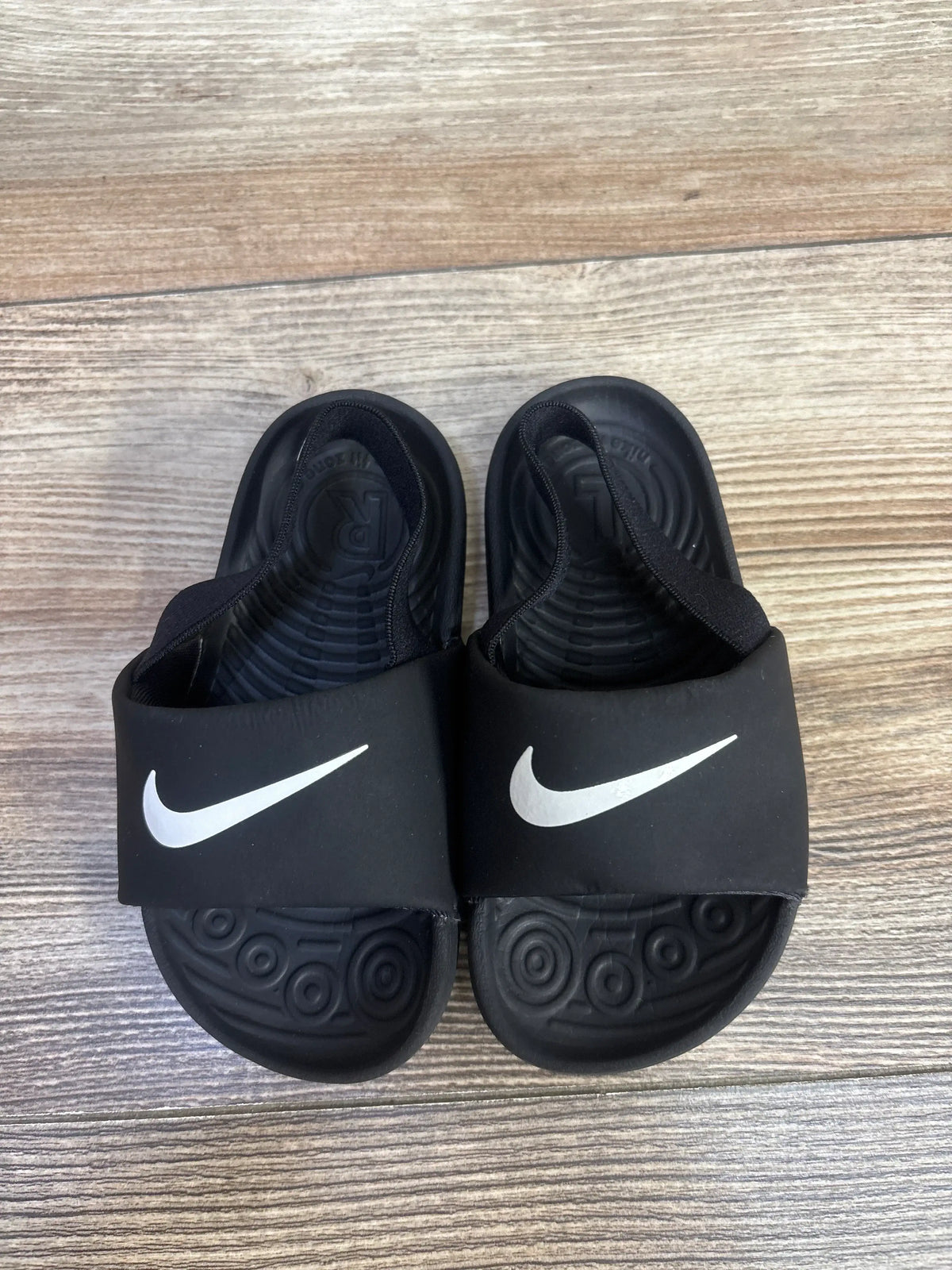 Top-down view of Nike Kawa slide sandals in black showing white Nike swooshes and textured footbeds on wood floor