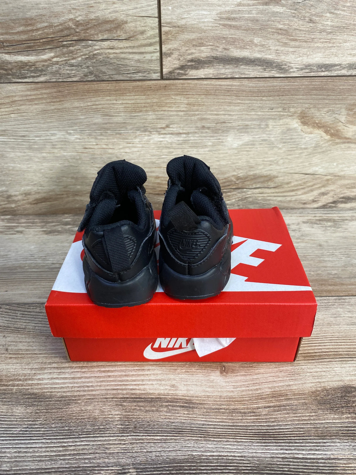 Rear view of Nike Air Max 90 EasyOn TD toddler sneakers in triple black showing heel pull tabs on red Nike box on wood floor