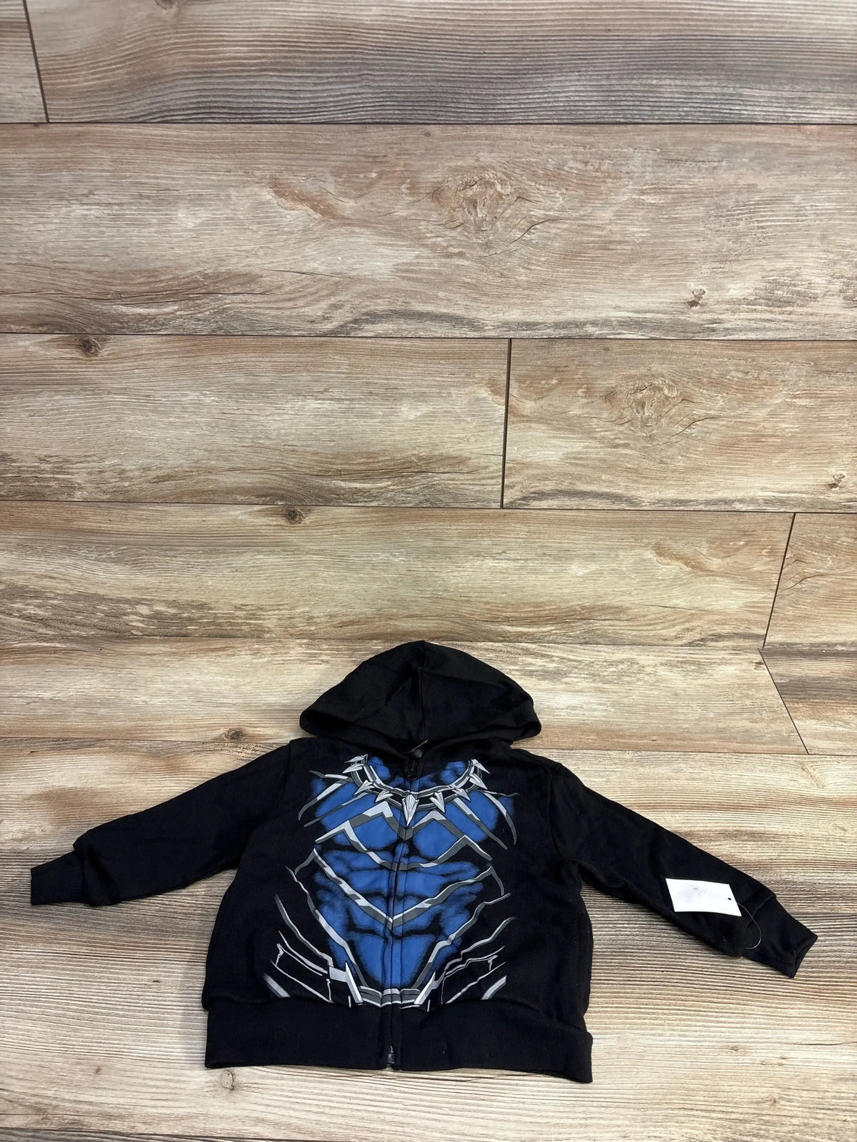 Kids black zip-up hoodie with blue superhero chest graphic, laid flat on wood floor