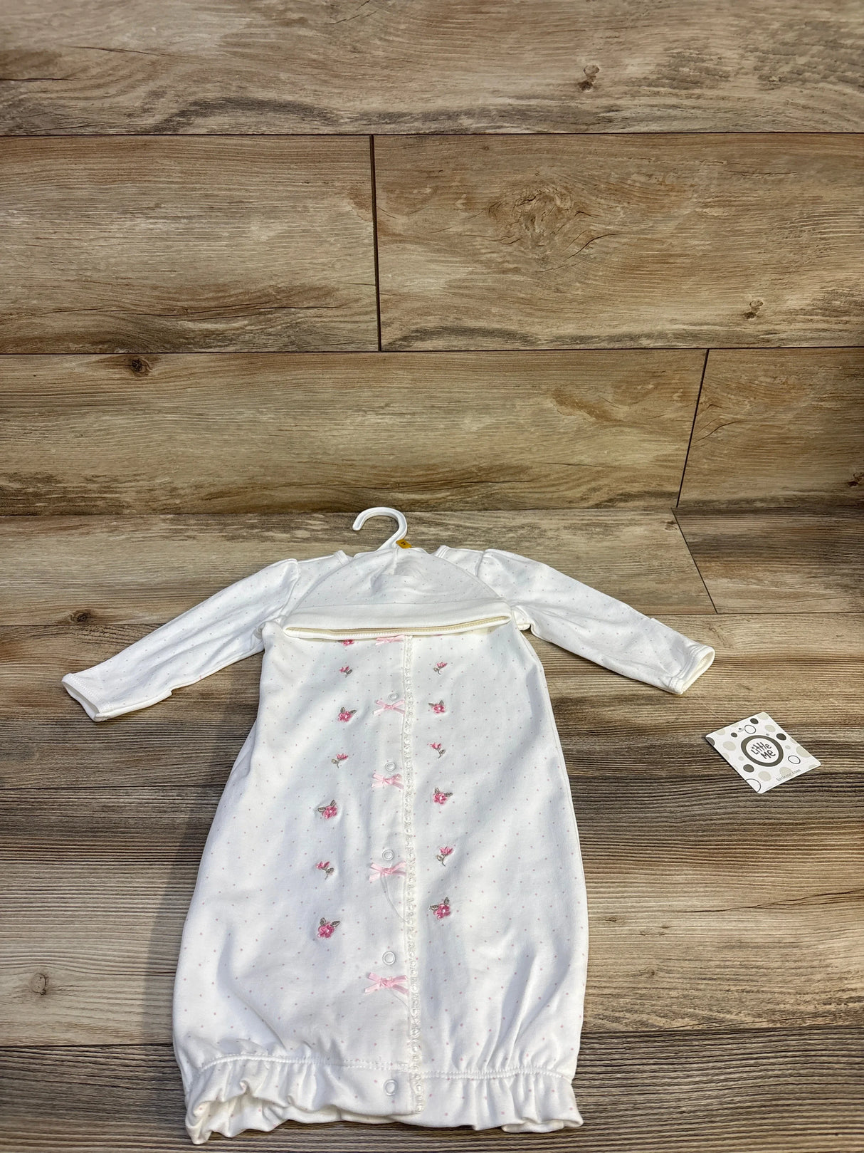 Front view of newborn white Little Me sleep gown and matching hat showcasing pink embroidered flowers down the front and snap closures on wood background