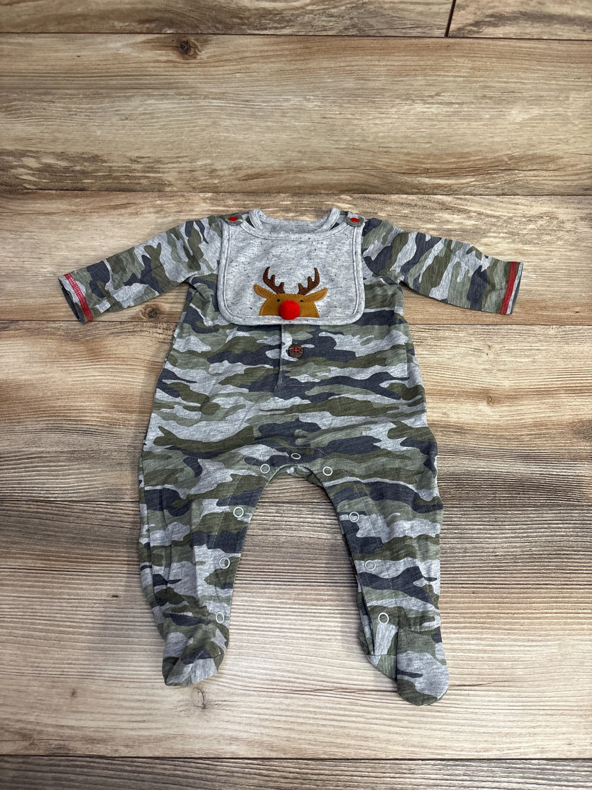 Baby camo footed romper with long sleeves and a reindeer bib featuring a red nose on wooden background