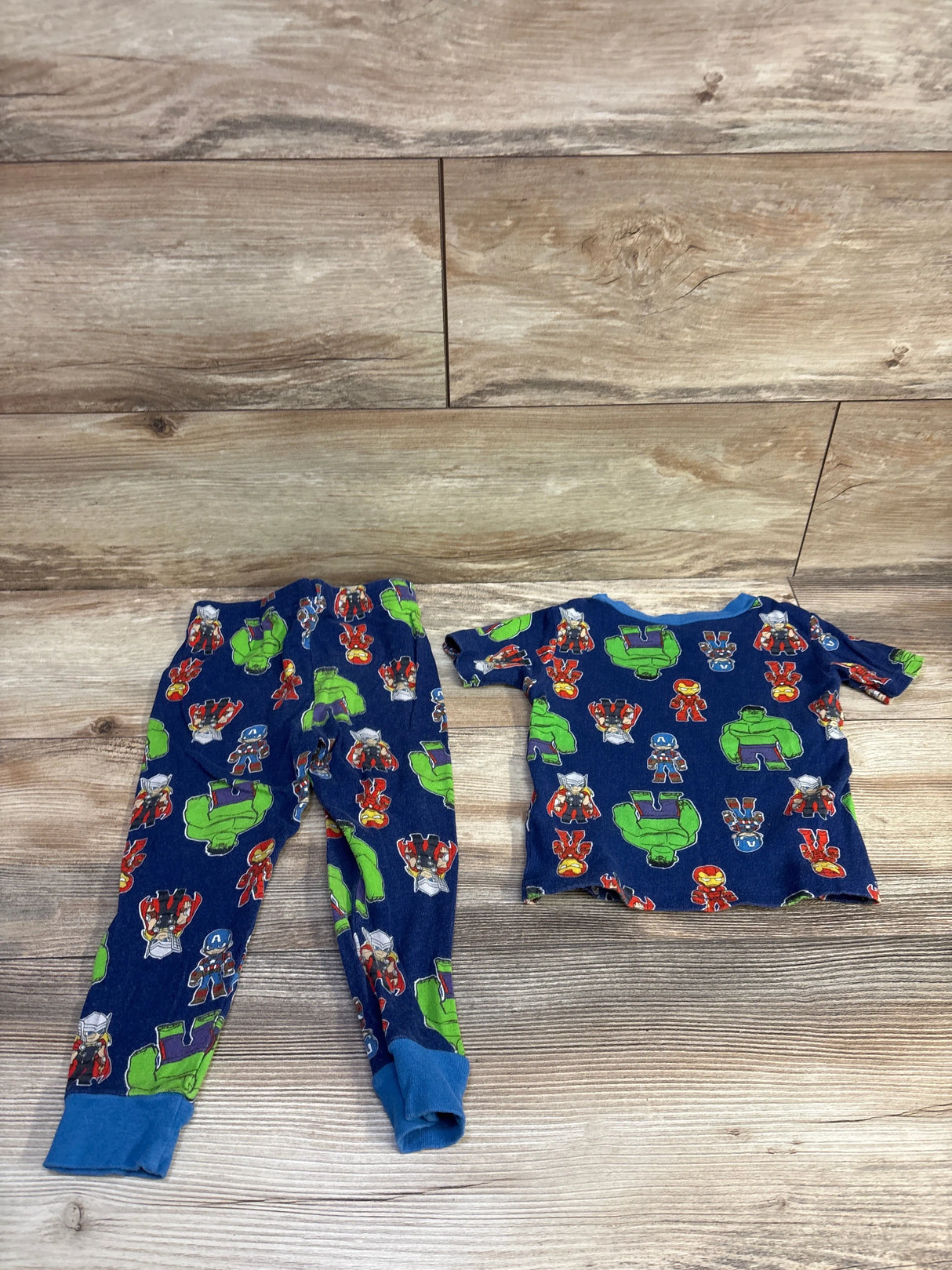 Flat-lay of Marvel Avengers 2-piece toddler pajama set showing navy blue top and cuffed pants with Hulk, Iron Man, Captain America and Thor prints on wood background