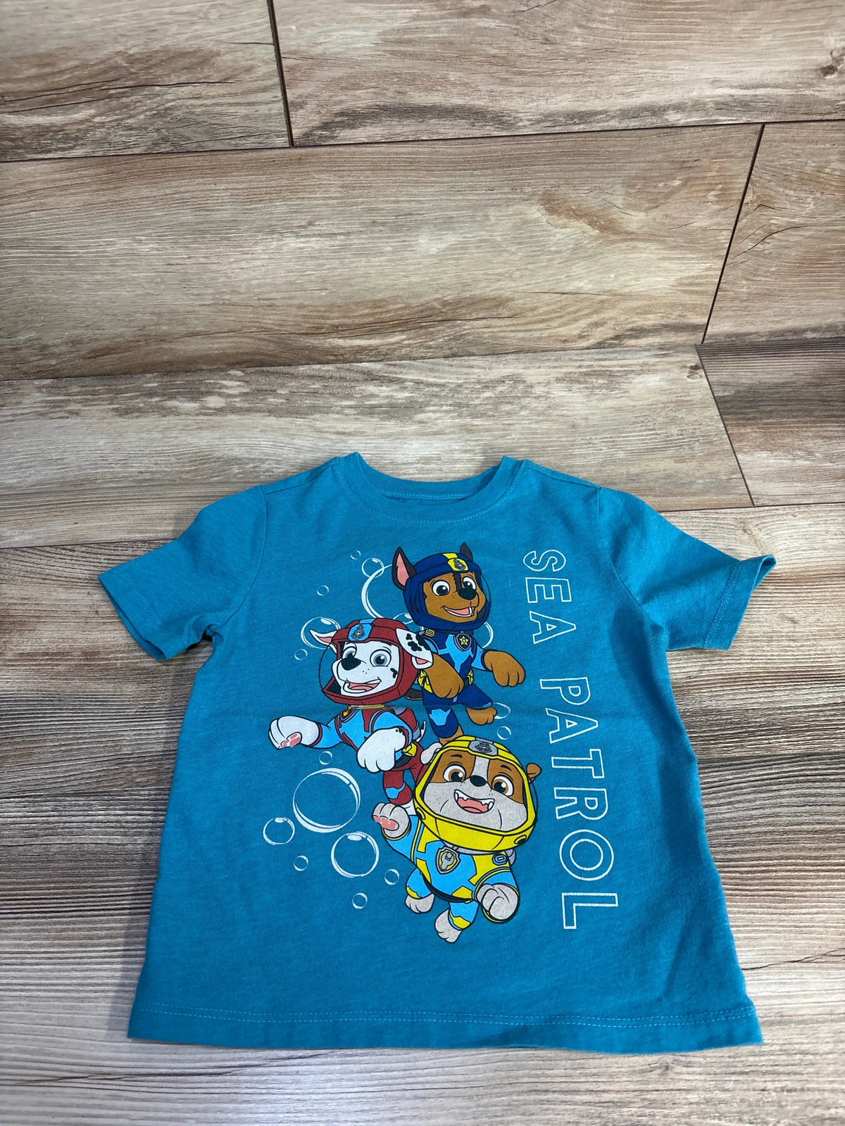 Front view of Old Navy PAW Patrol shirt showcasing blue Sea Patrol character graphic with a wood-look background