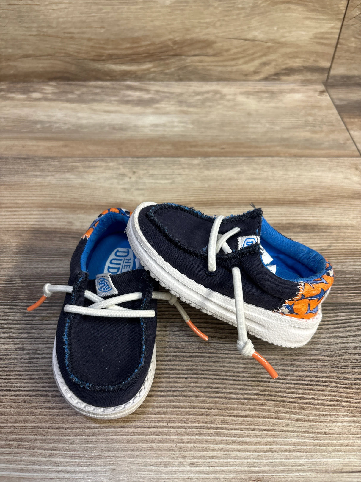 Three-quarter view of Hey Dude Wally toddler navy shoes with orange shark-print heel, white elastic laces and white sole on wood background