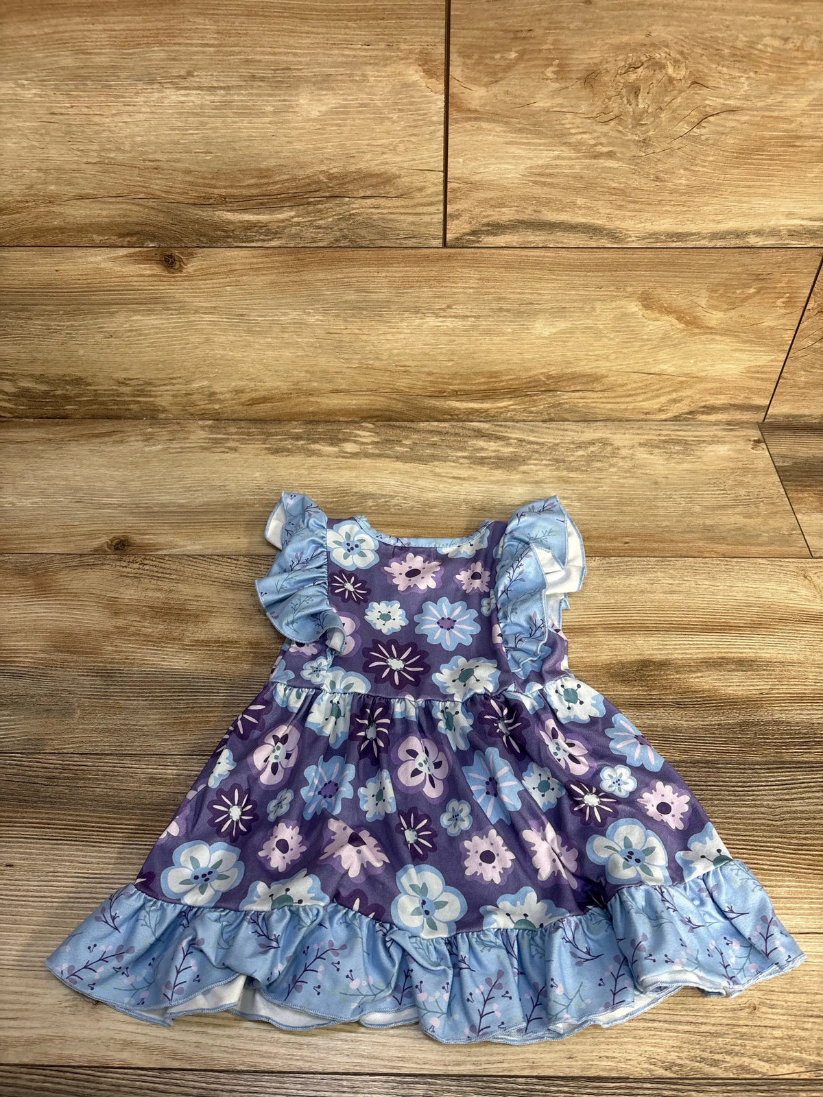 Front view of Pete + Lucy blue floral dress showcasing ruffled sleeves and hem on a wood floor