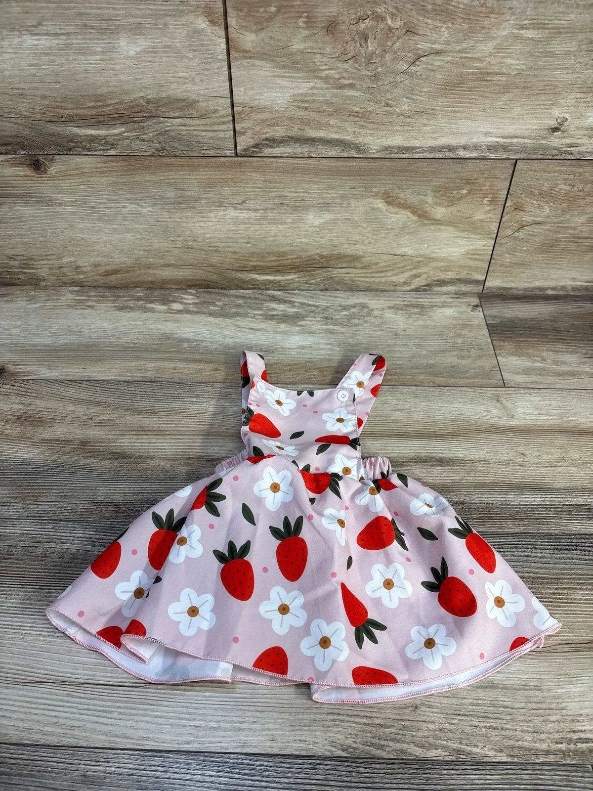 Flat lay of pink strawberry-print skirtall with button shoulder straps, gathered elastic waist, and full skirt with white flowers on wood floor