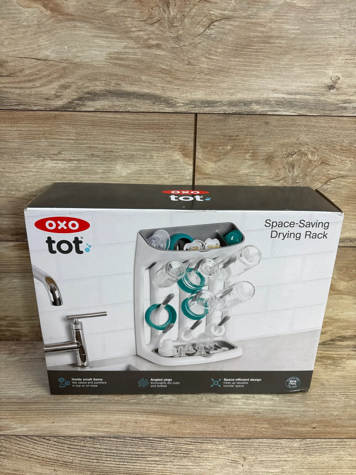 Front view of OXO Tot space-saving drying rack box showing the rack with angled bottle pegs and accessory tray on wood backdrop