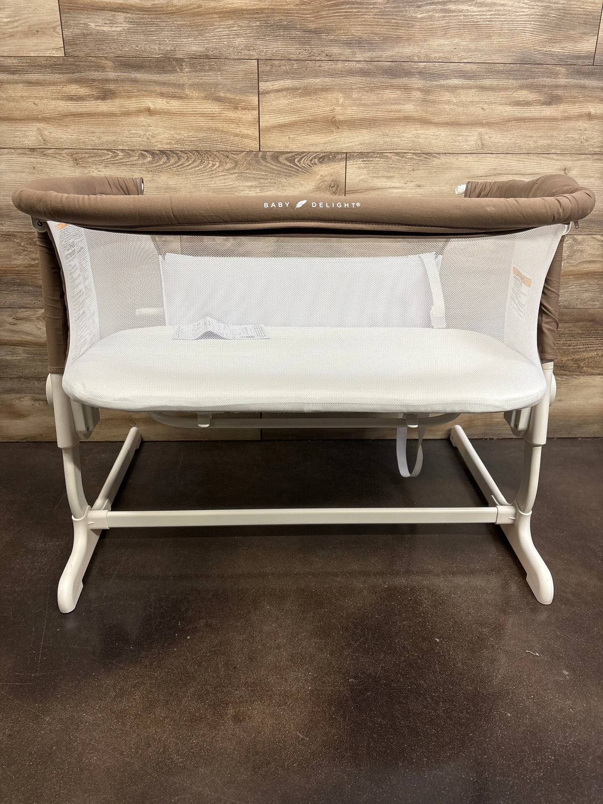 Front view of Beside Me Dreamer bedside bassinet showcasing mocha padded top rail, mesh sides, and white frame against wood-paneled wall